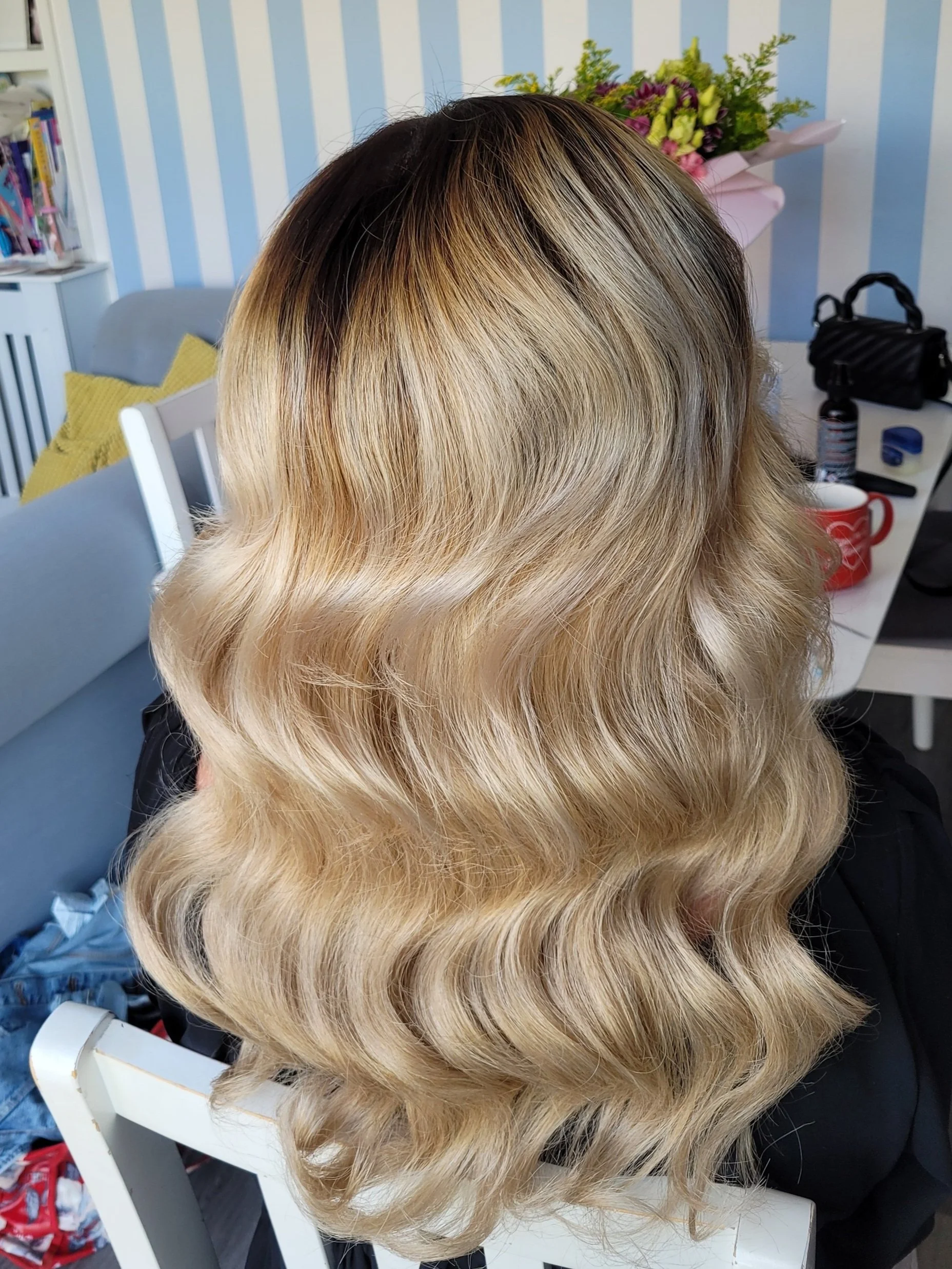 Close-up of long, wavy, blonde hair with dark roots, styled over the back of a white chair, in a cozy room setting with floral decorations and a striped wall.