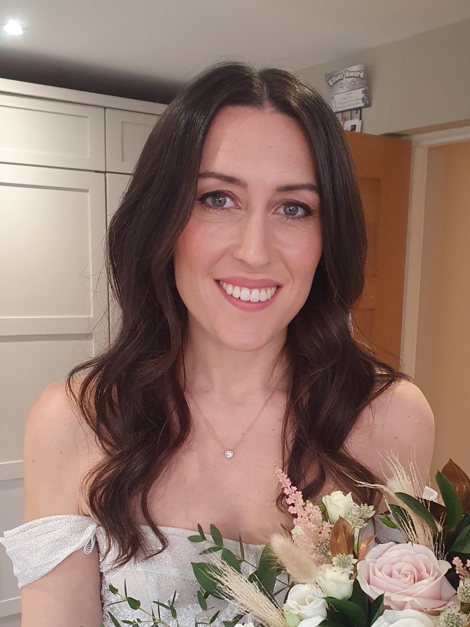 Smiling woman with long brown hair, wearing a white dress and holding a bouquet with pink and white flowers.