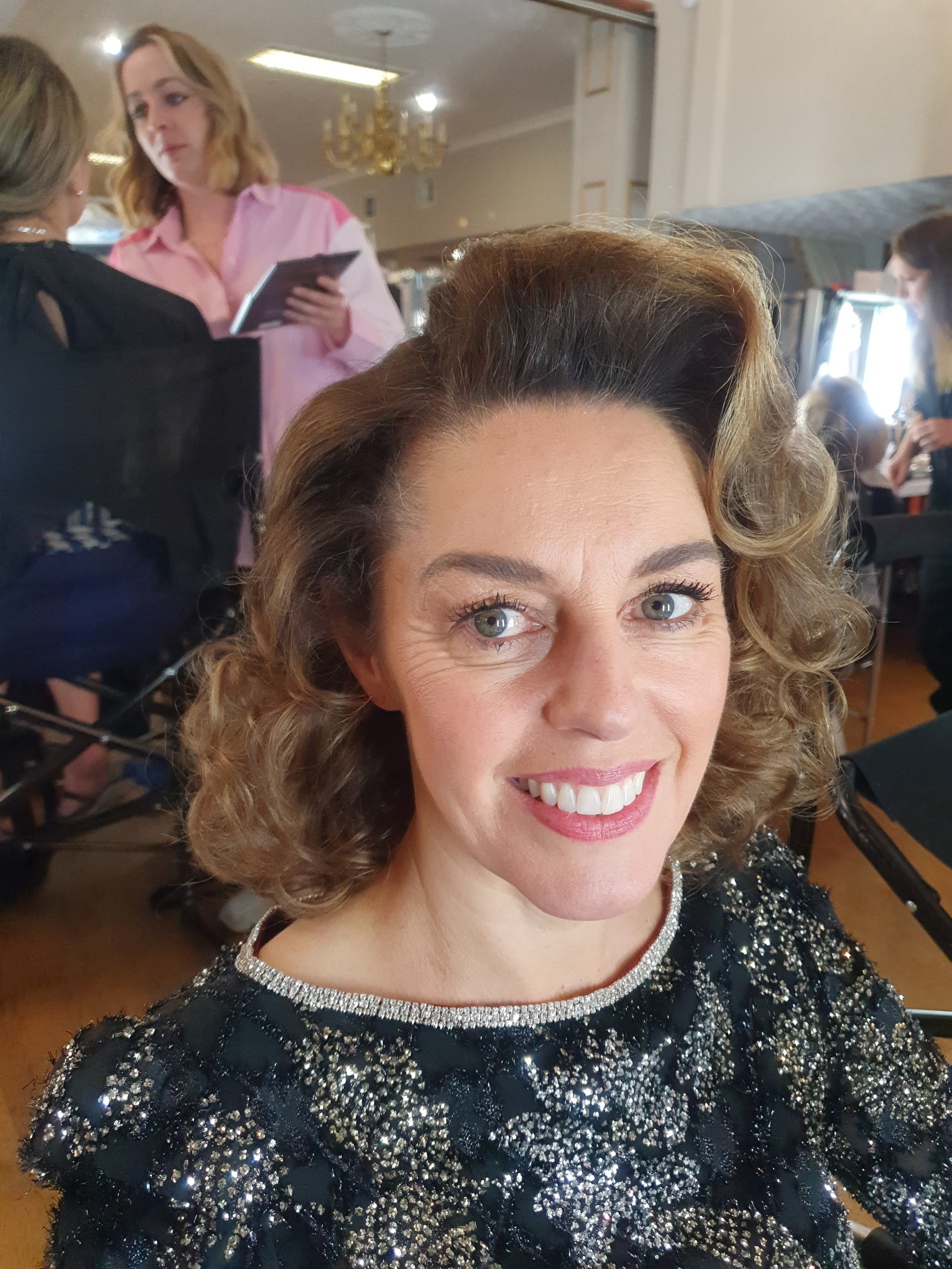 Woman with curly hair and sequined dress smiling indoors, with people in the background.