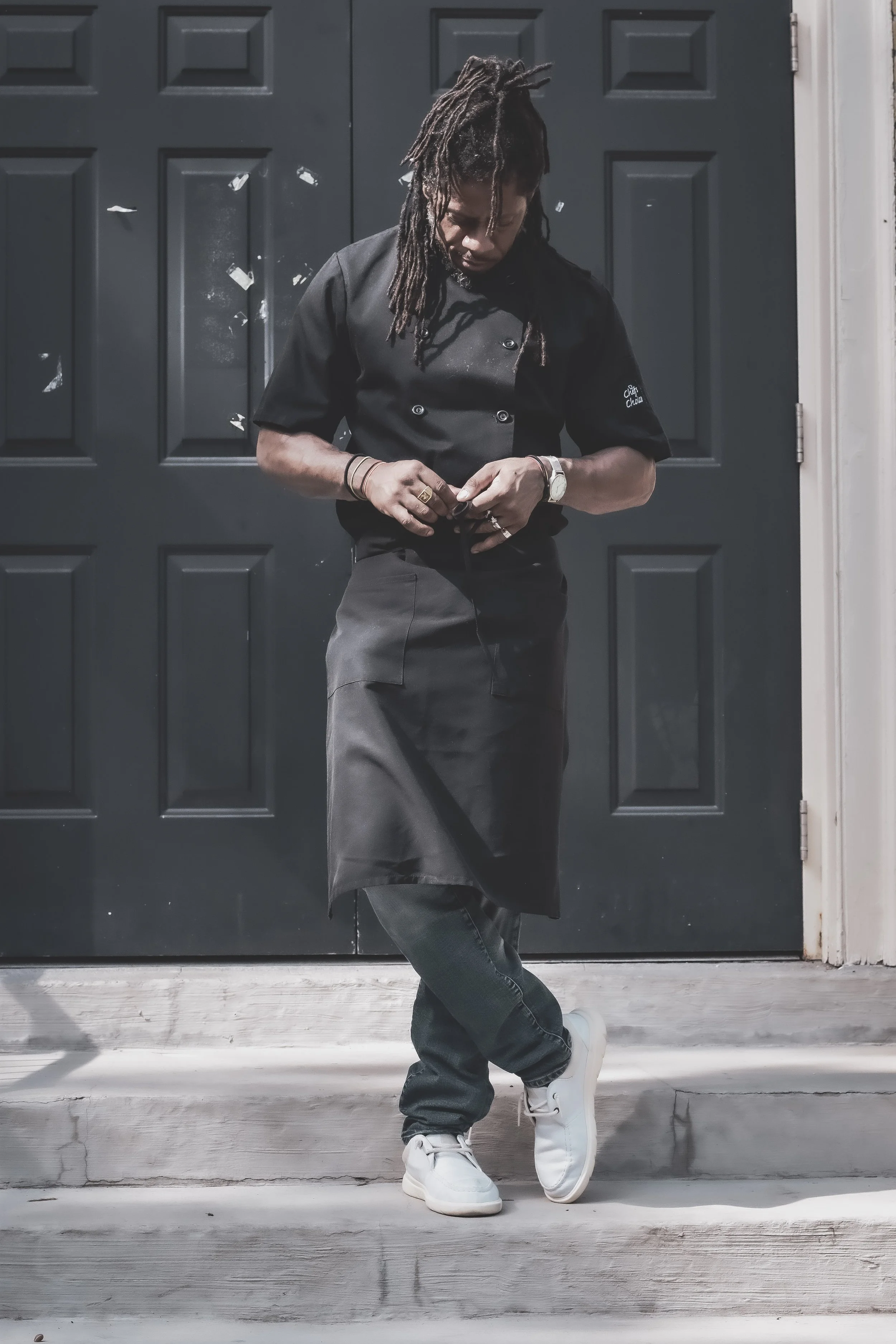 Man standing in chef uniform with black apron and white sneakers, in front of dark doors.