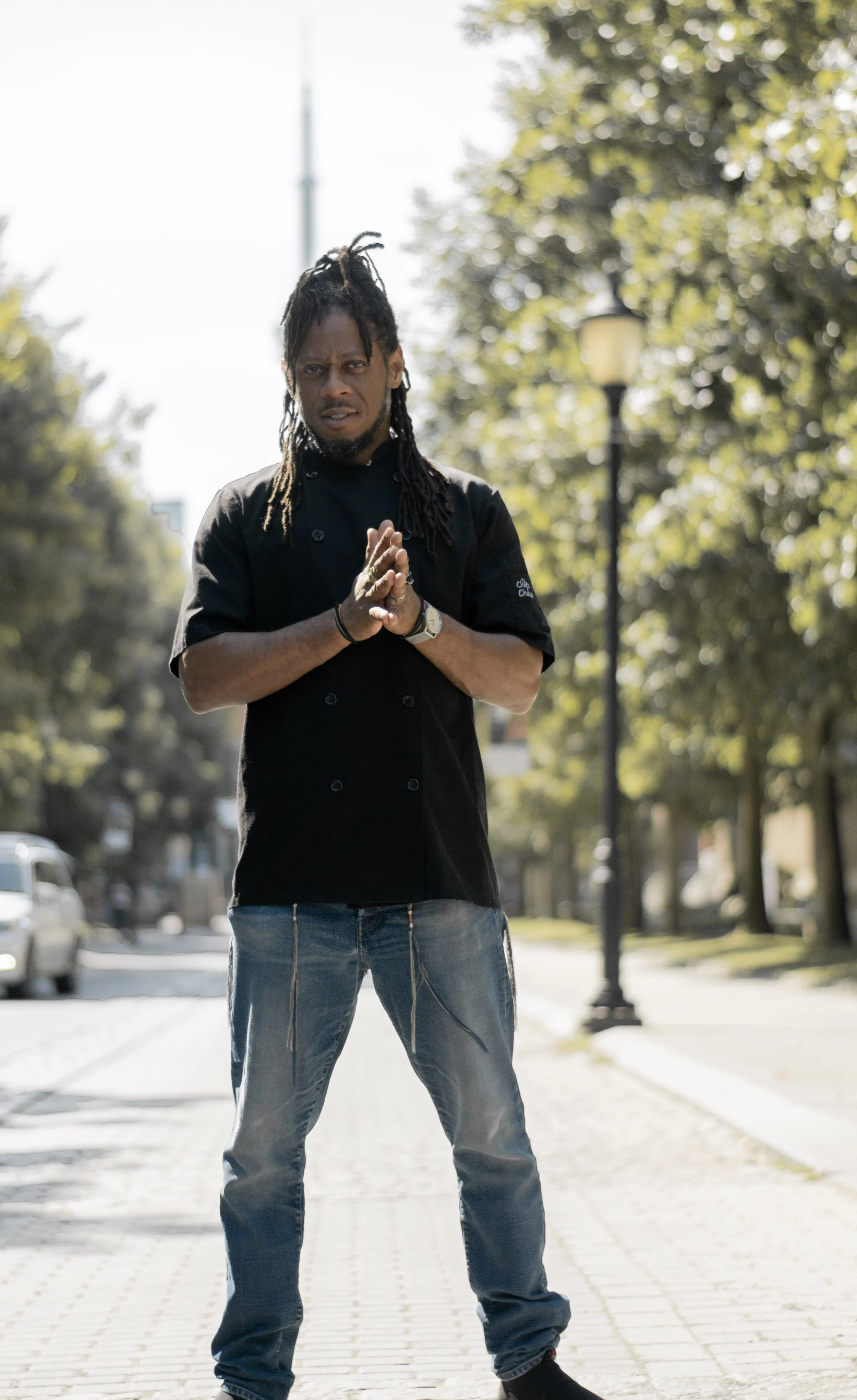 A person with dreadlocks wearing a black chef's coat and jeans standing outdoors on a brick pathway with trees and a streetlamp in the background.