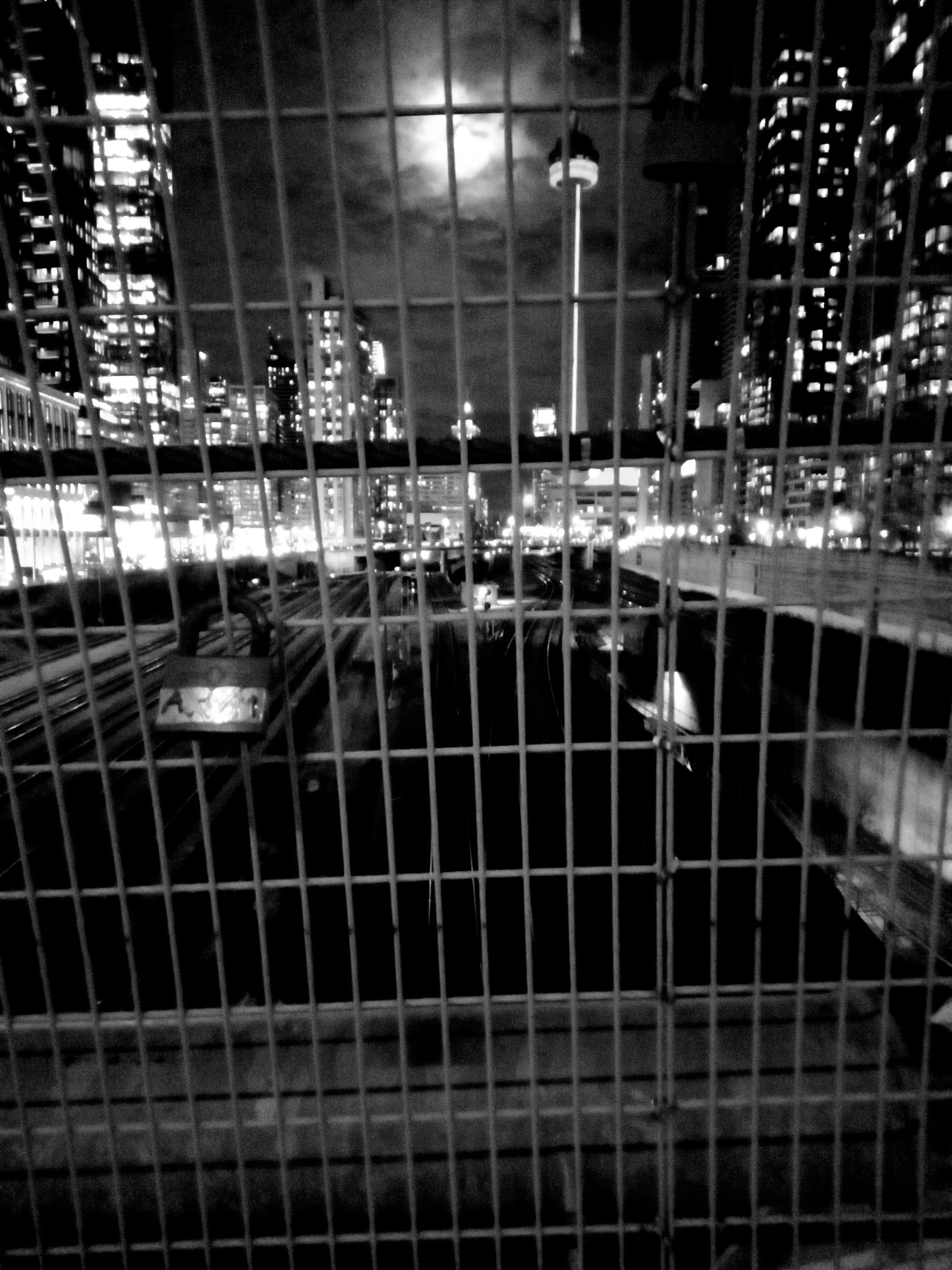 Black and white photo of a cityscape behind metal fencing, featuring tall buildings and an illuminated tower at night.