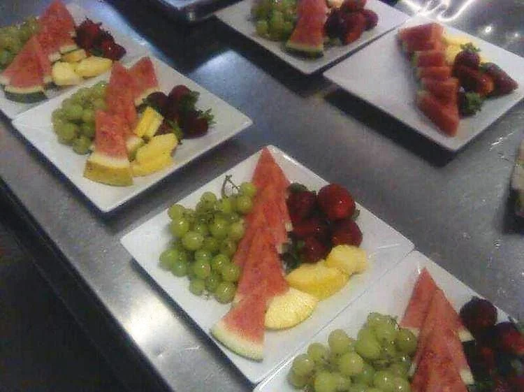 Plates with sliced watermelon, grapes, strawberries, and pineapple on a metal surface.