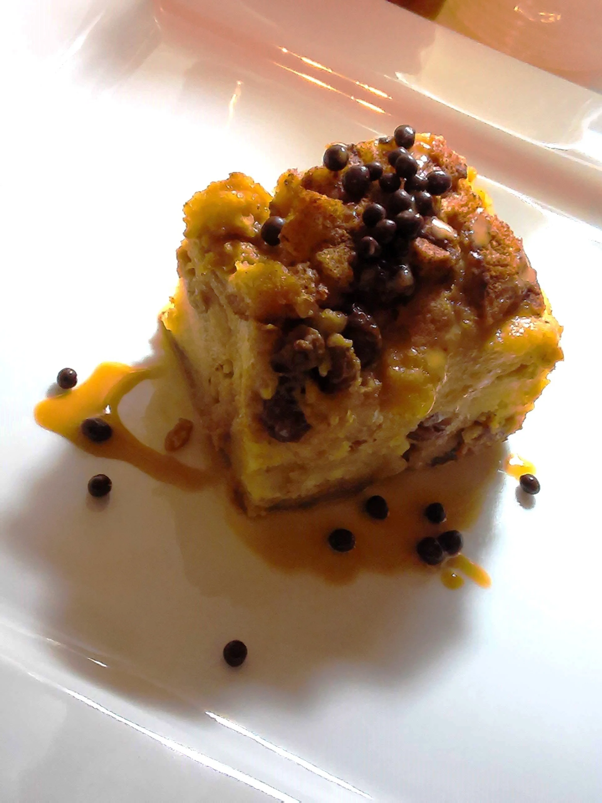 A piece of bread pudding garnished with chocolate covered Quinoa puffs and burnt caramel & Bourbon sauce on a white plate.