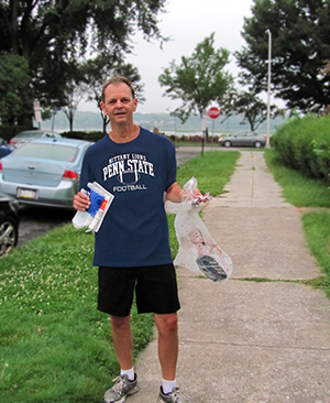 Dan Miller standing on sidewalk holding a plastic bag with items and a water bottle, wearing a Penn State football T-shirt, shorts, and sneakers, near parked cars and trees.