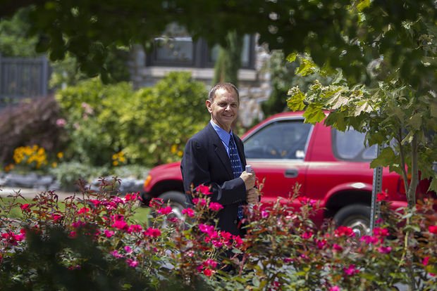 A man in a suit holding a drink, standing outdoors behind pink flowers and green foliage, with a red vehicle and a residential building in the background.