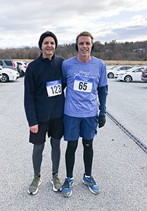 Two runners, a woman and Dan Miller, standing together in a parking lot after a race. They are wearing race bibs with numbers 123 and 65, and athletic clothing. Both are smiling.