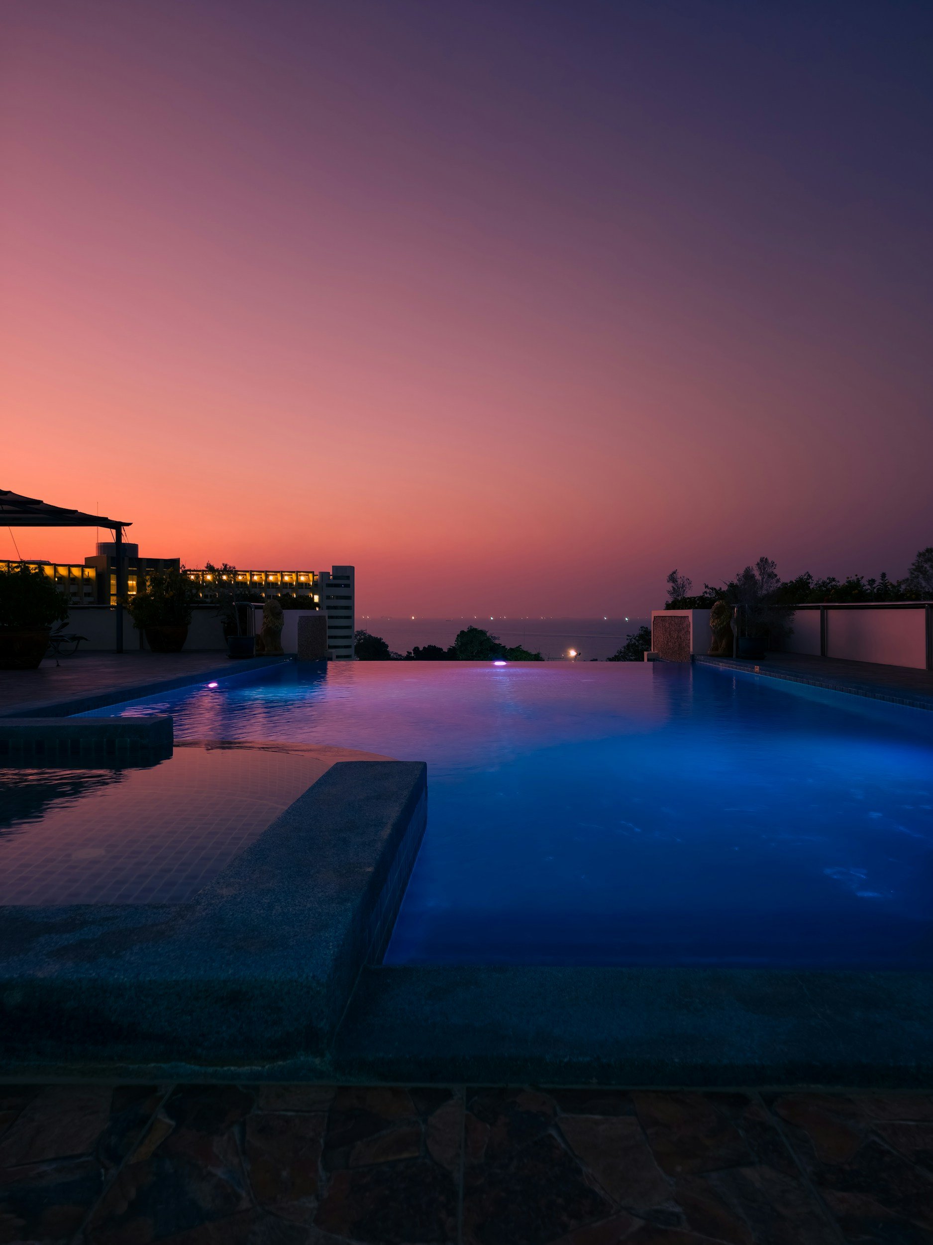 A rooftop infinity pool overlooking a sunset with a pink and purple sky, distant city lights, and trees on the horizon.