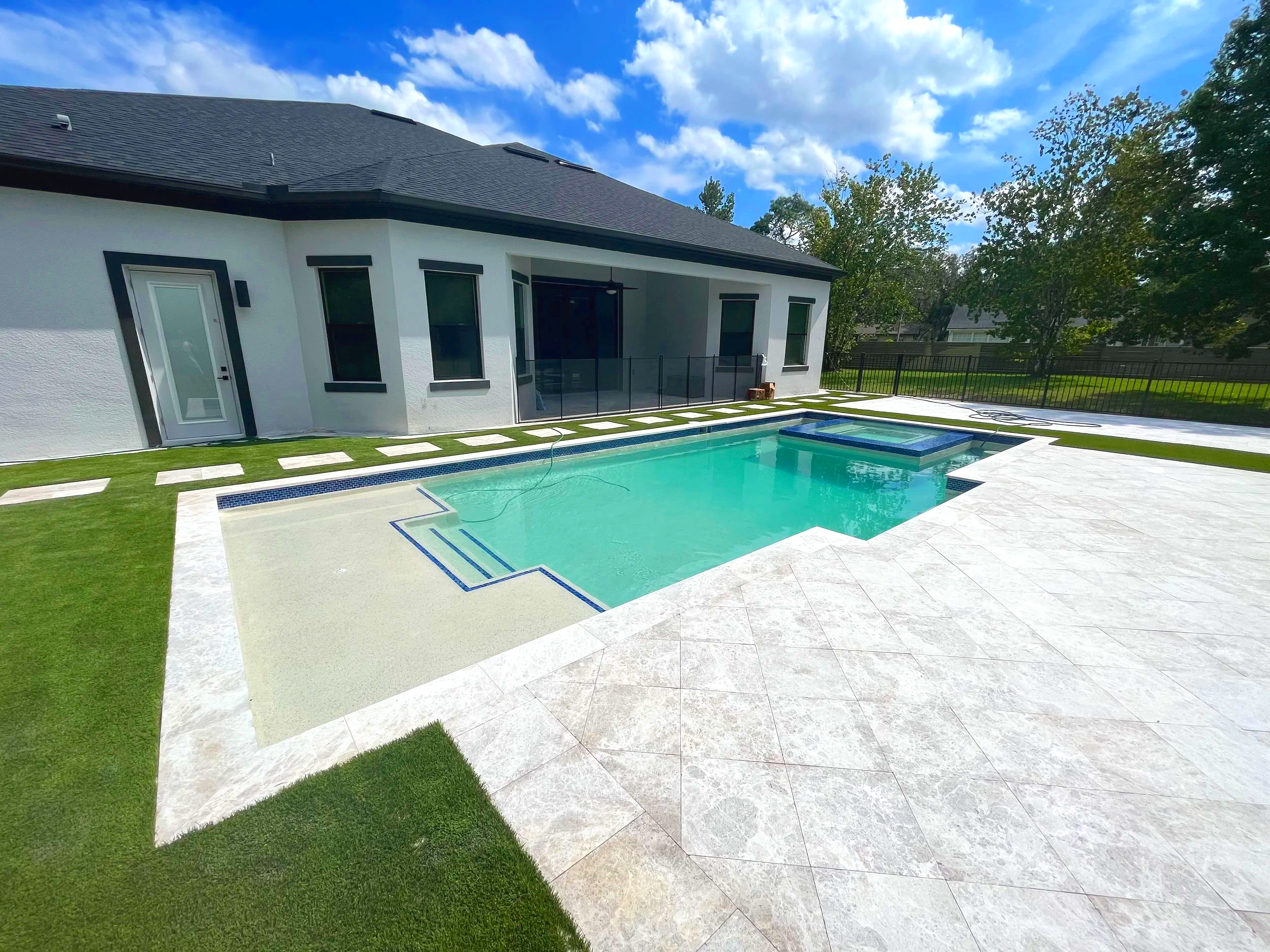 Backyard with a swimming pool and spa, adjacent to a modern white house with black trim, green lawn, and trees under a partly cloudy sky.