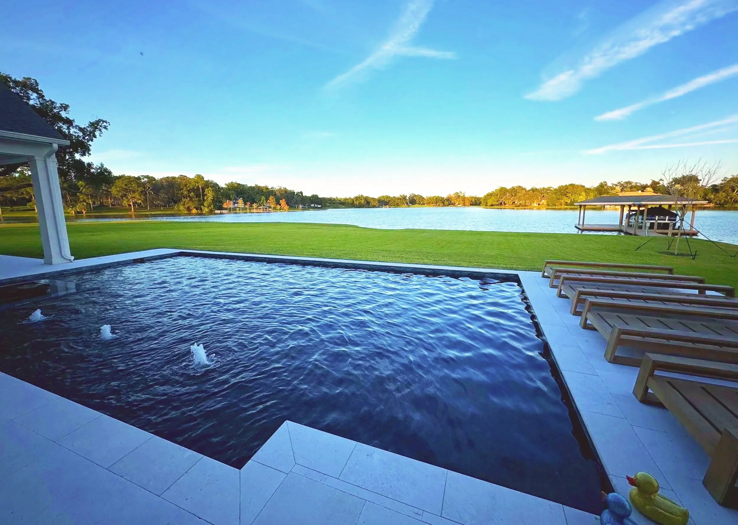 Outdoor view of a waterfront scene with a pool in the foreground, overlooking a lake with a grassy lawn and trees in the background. There are wooden lounge chairs on the patio, and a covered boat dock extends over the water.