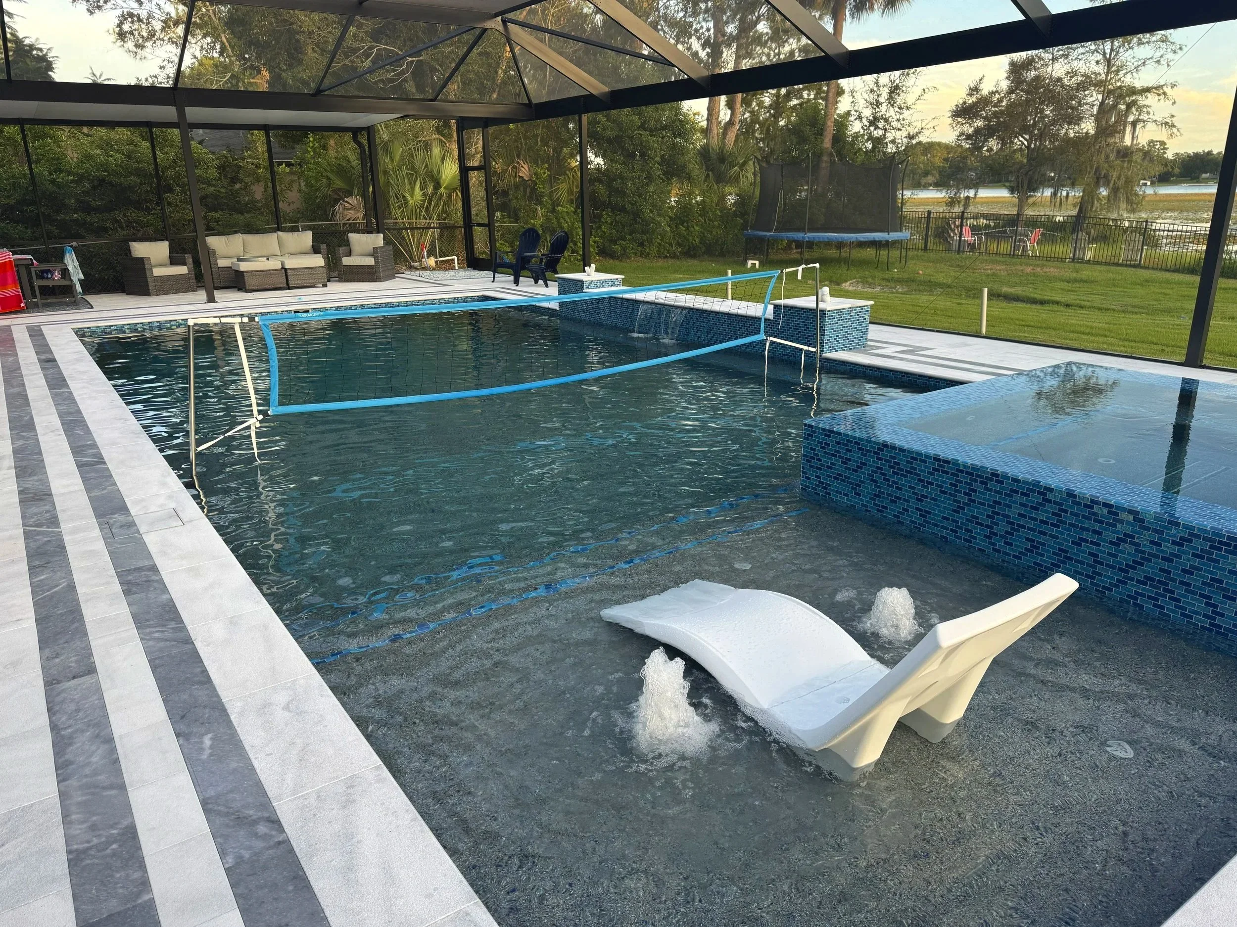 Backyard swimming pool enclosed by a screened porch, with lounge chairs, outdoor seating, a trampoline, and a grassy yard in the background.