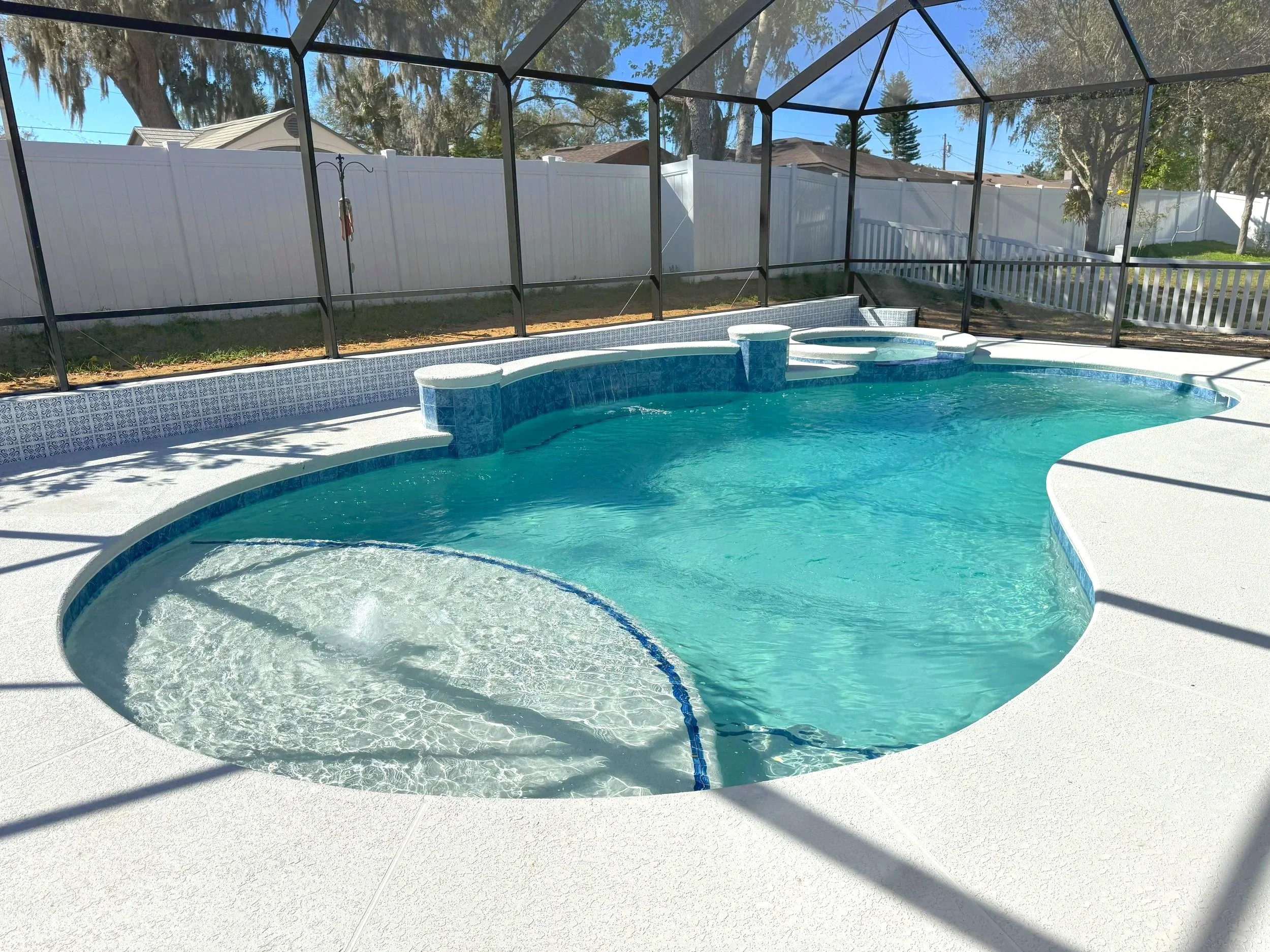 A backyard pool enclosed by a black metal and screen enclosure, with a white privacy fence, trees, and houses in the background, under a clear blue sky.