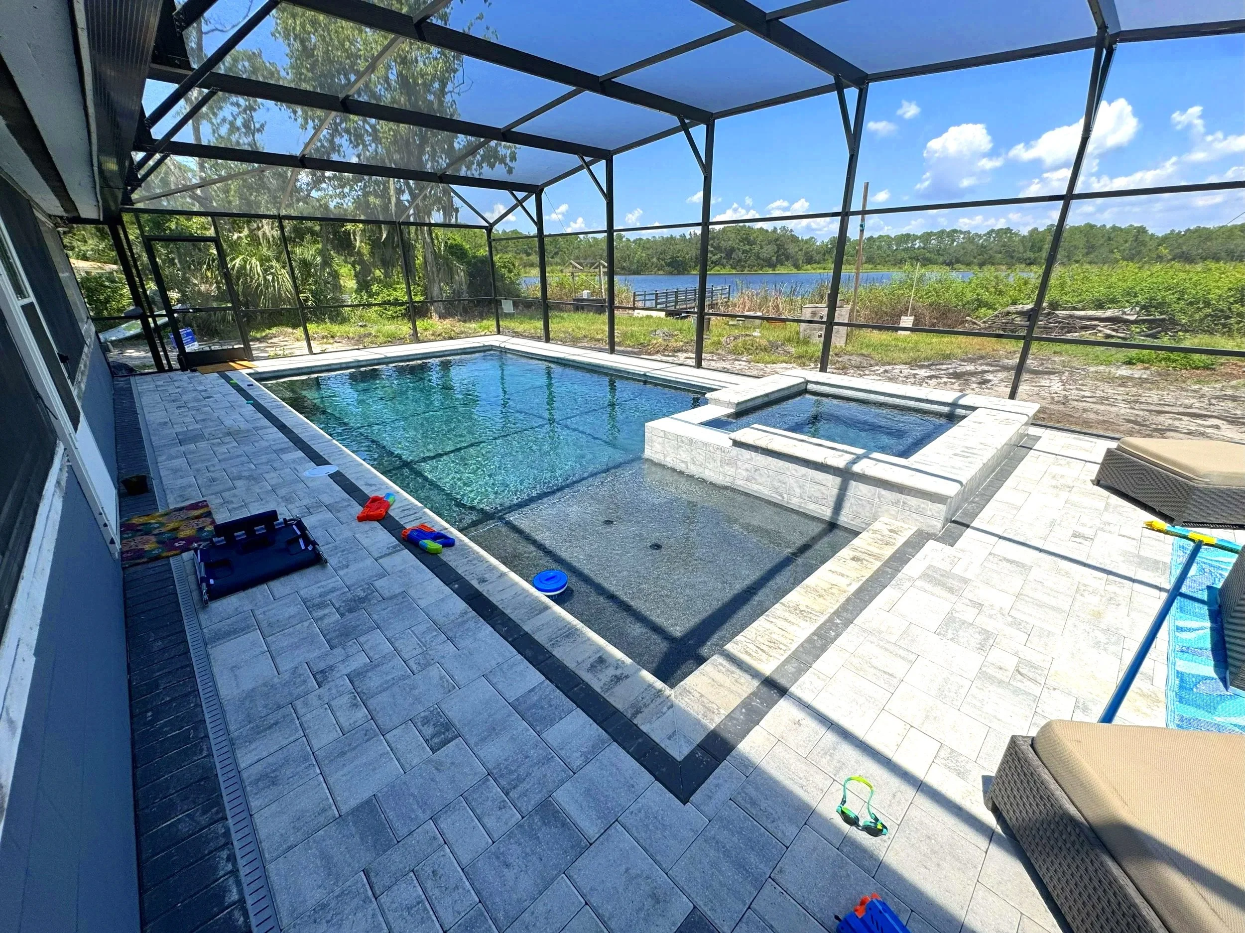 Screened-in backyard pool area with a lake view, featuring a rectangular pool with a small attached hot tub, outdoor lounge chairs, and scattered children's toys, under a blue sky with clouds.