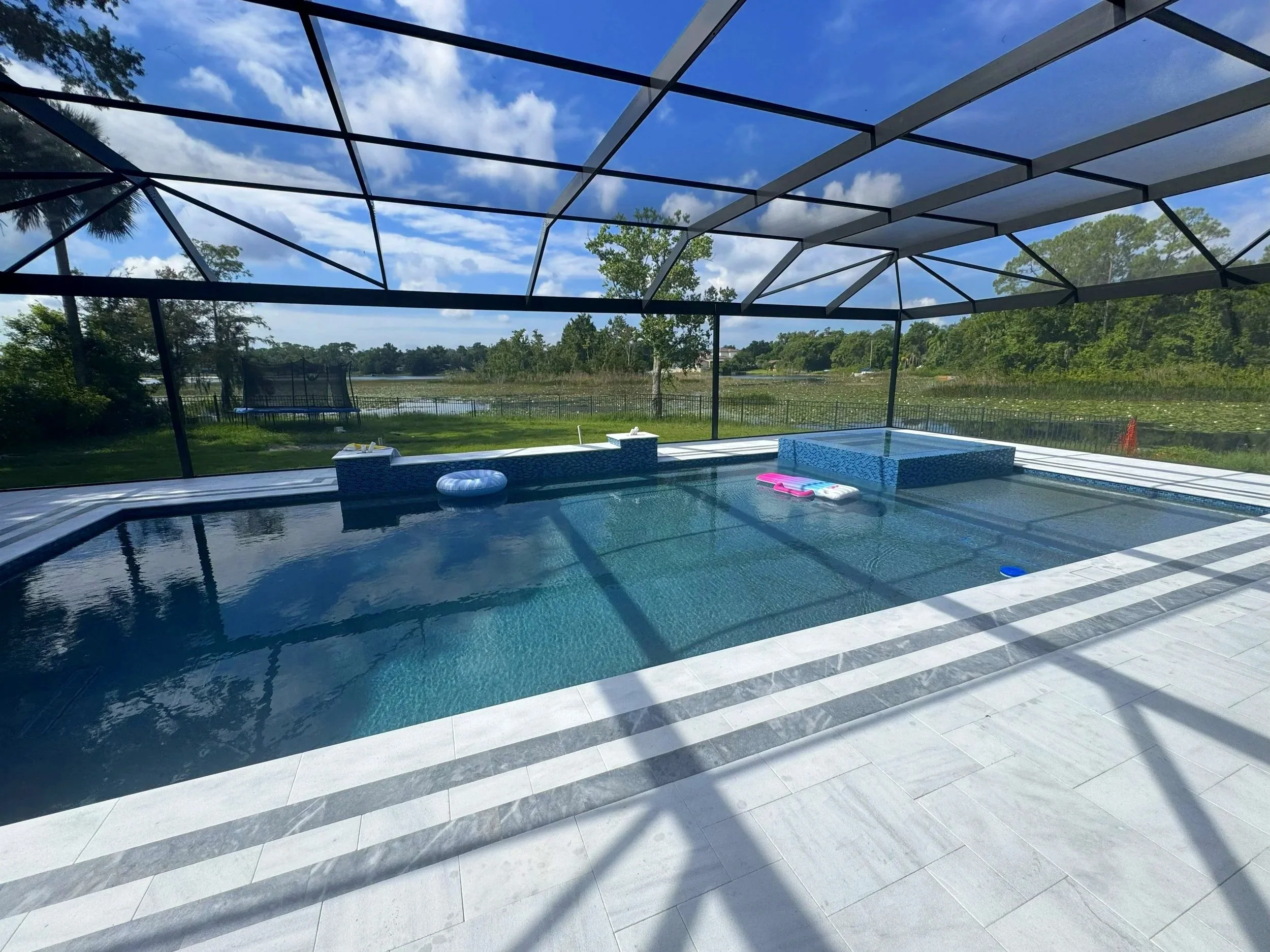 Enclosed backyard swimming pool area with a screened roof, surrounded by white tiles, overlooking a lake and greenery.