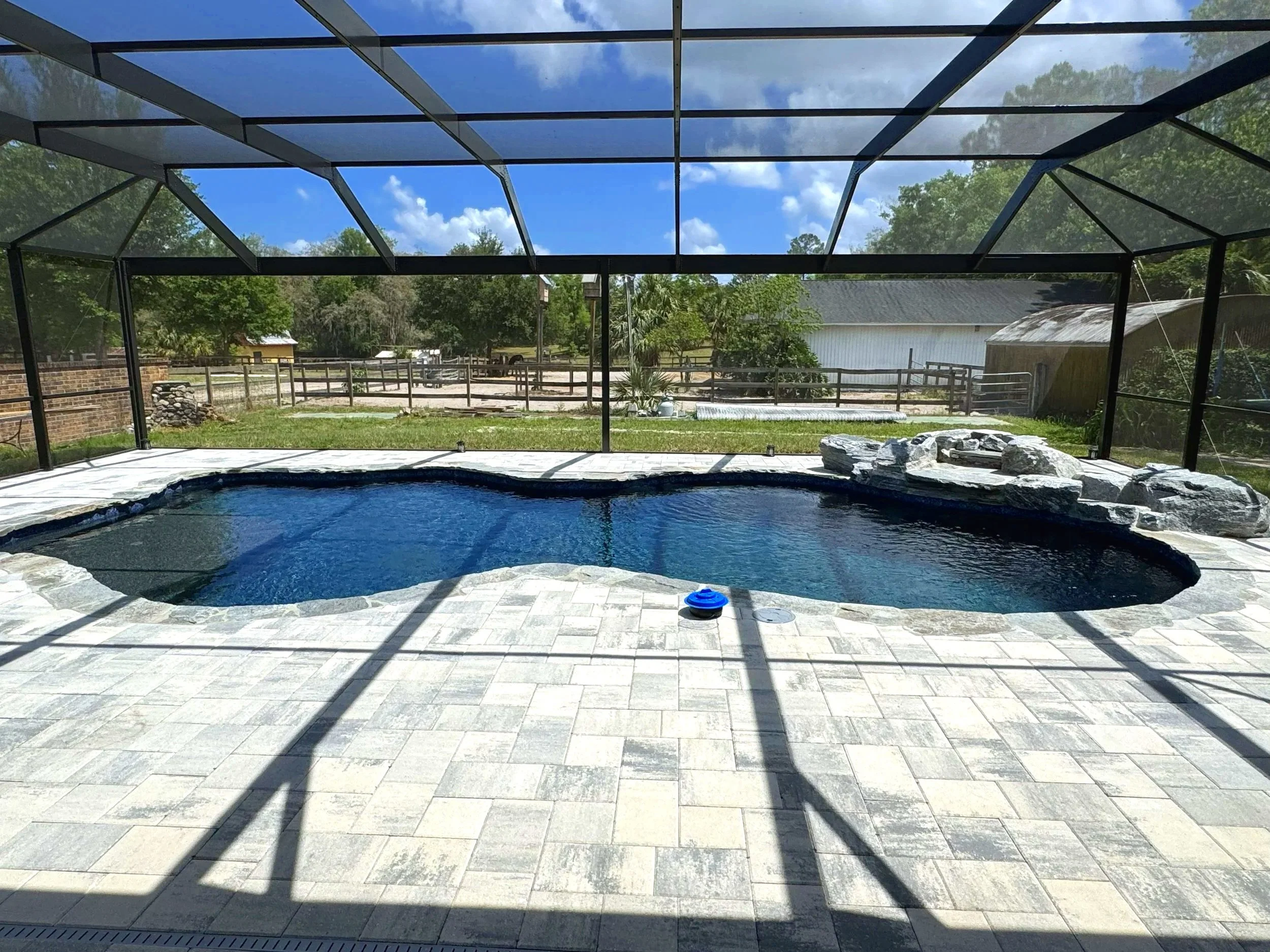 Enclosed backyard pool area with a dark blue freeform swimming pool, stone waterfall feature on the right, and a screened enclosure. The background shows a grassy yard, trees, and a white building with a sloped roof under a partly cloudy sky.
