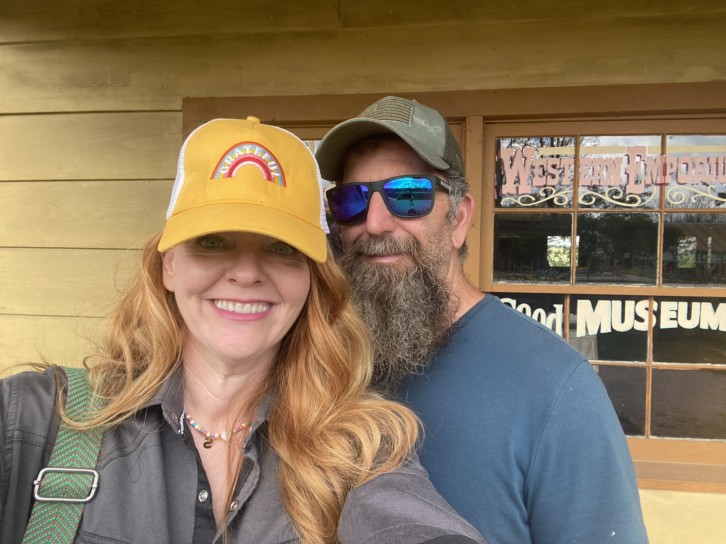 A smiling woman with red hair wearing a yellow cap and a man with a beard, sunglasses, and a green cap taking a selfie outside a building with yellow walls and a window that has signs reading 'Western Emporium' and 'Good Museum'.