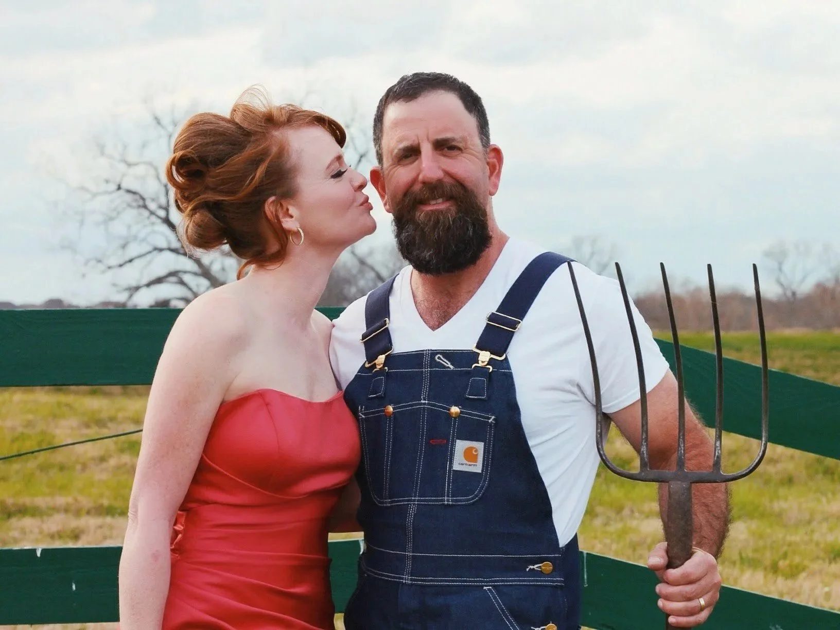 A woman in a strapless red dress kisses a man with a beard, wearing a white t-shirt and denim overalls, holding a pitchfork outdoors.