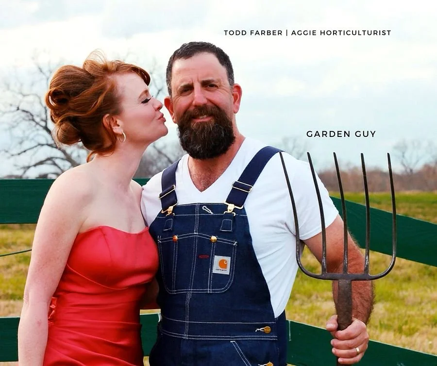 Man with a beard wearing overalls holding a pitchfork, woman with red hair kissing his cheek, outdoor setting, fields and trees in background.
