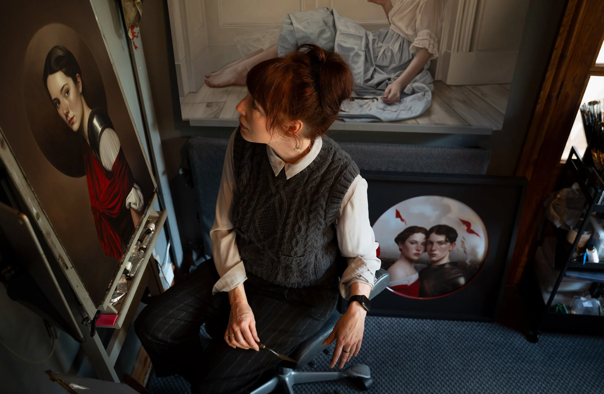 A woman with red hair, wearing a white shirt and gray knit vest, sitting in an art studio looking at a portrait of a woman dressed in a kimono with a red sash, in front of her.