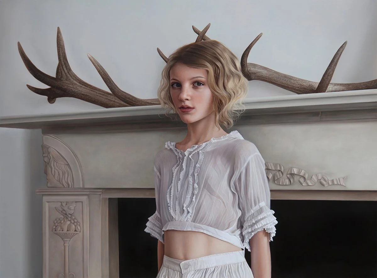 Young woman with wavy blonde hair standing indoors in front of a fireplace decorated with antlers. She is wearing a sheer, white, ruffled crop top and matching high-waisted shorts.