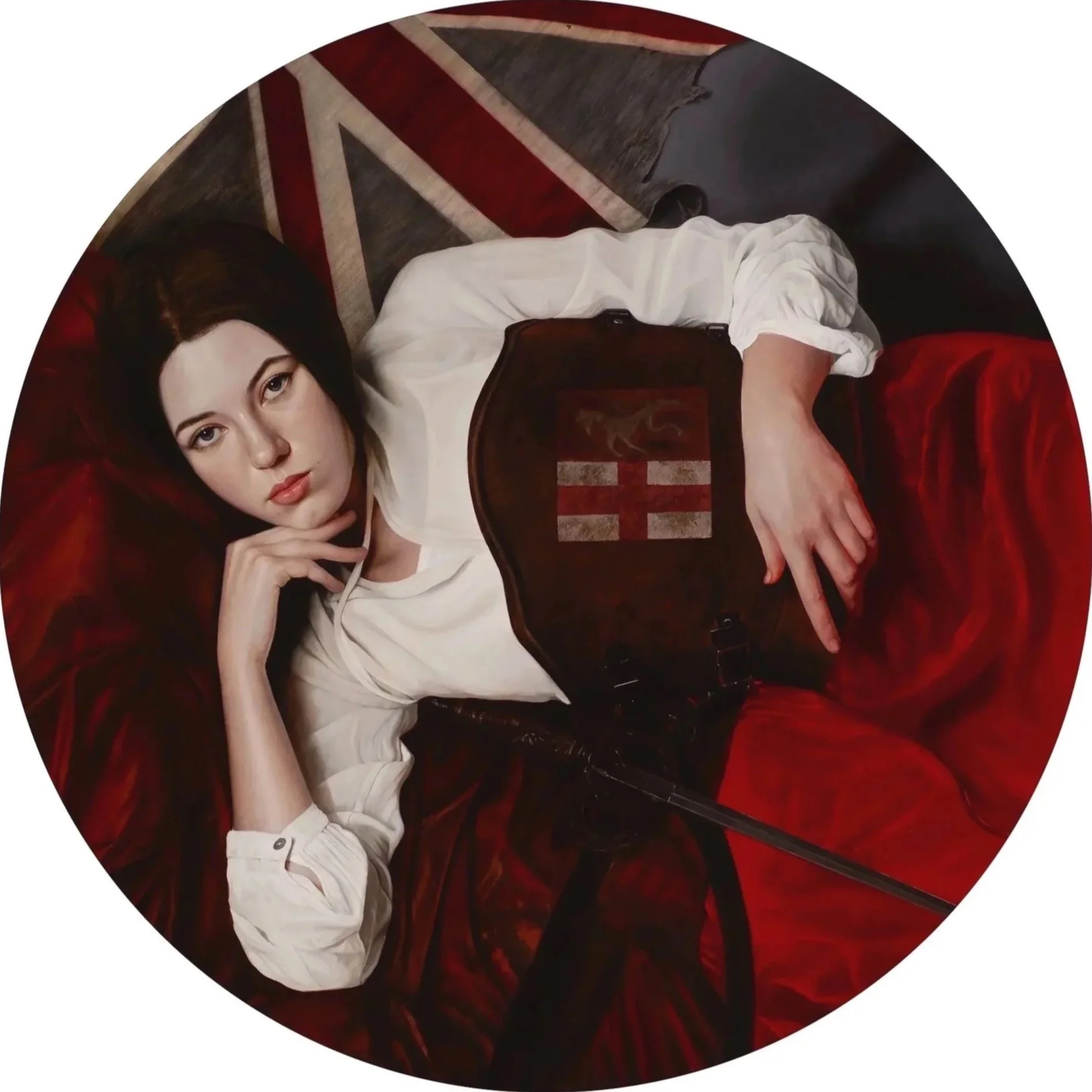 A woman with dark hair and fair skin lying on a red velvet surface, wearing a white blouse and a dark leather apron with a medieval English cross emblem. She has a relaxed pose, resting her head on her hand and looking at the camera.