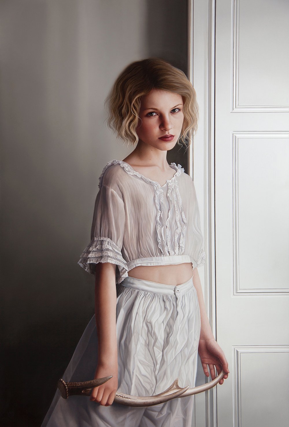 A young woman with wavy blonde hair and a serious expression, wearing a sheer, vintage-inspired white blouse with ruffles and white high-waisted pants, standing indoors near a white paneled door, holding a curved sword in her right hand.