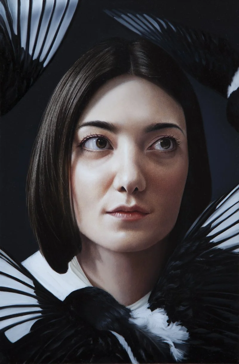 A woman with short dark hair and brown eyes is looking upwards. She is surrounded by black and white feathers that frame her face against a dark background.