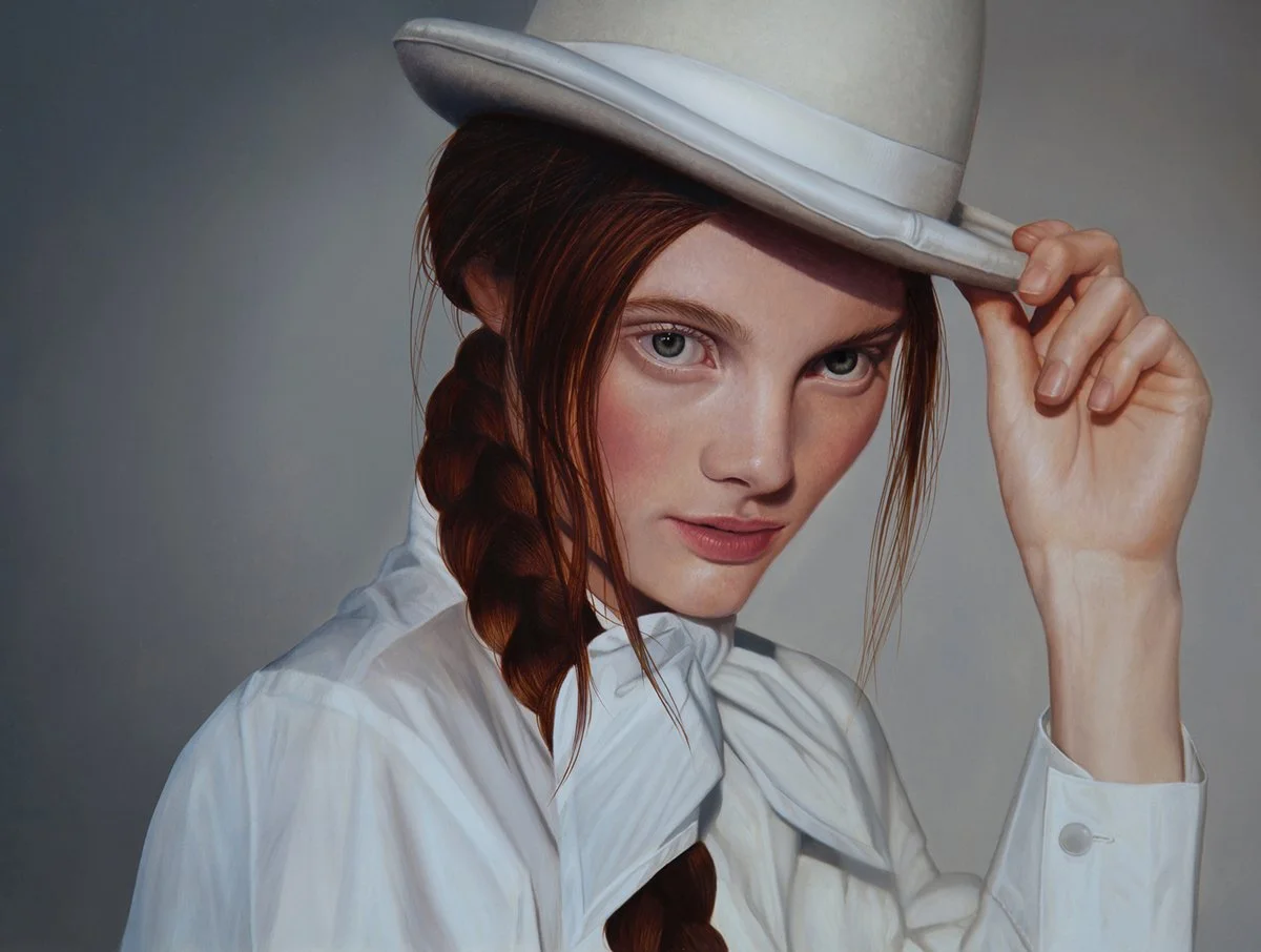 A woman with long red hair styled in a braid, wearing a white shirt and a light-colored hat, looking directly at the camera. The woman is holding the brim of her hat with her right hand.