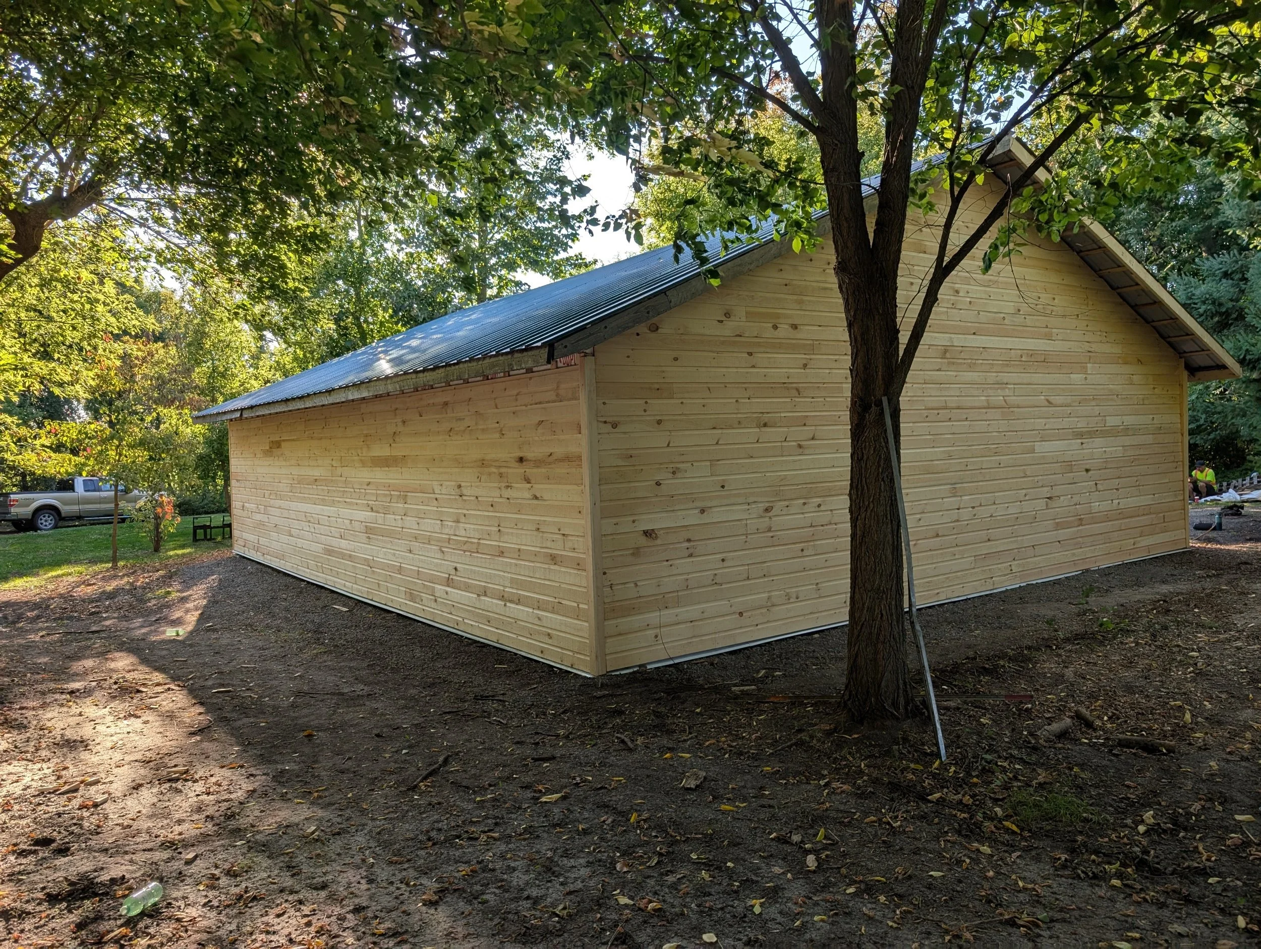 A wooden building under construction with new horizontal wooden siding and a sloped metal roof, surrounded by trees and a dirt ground.