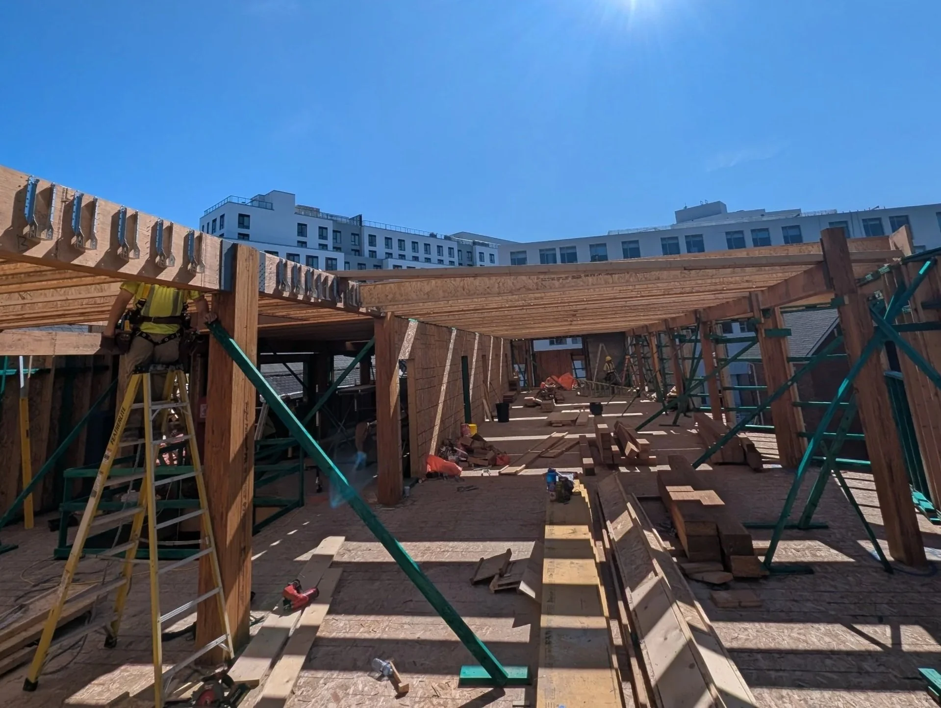 Construction workers building a wooden structure outdoors under a clear blue sky with modern white buildings in the background.