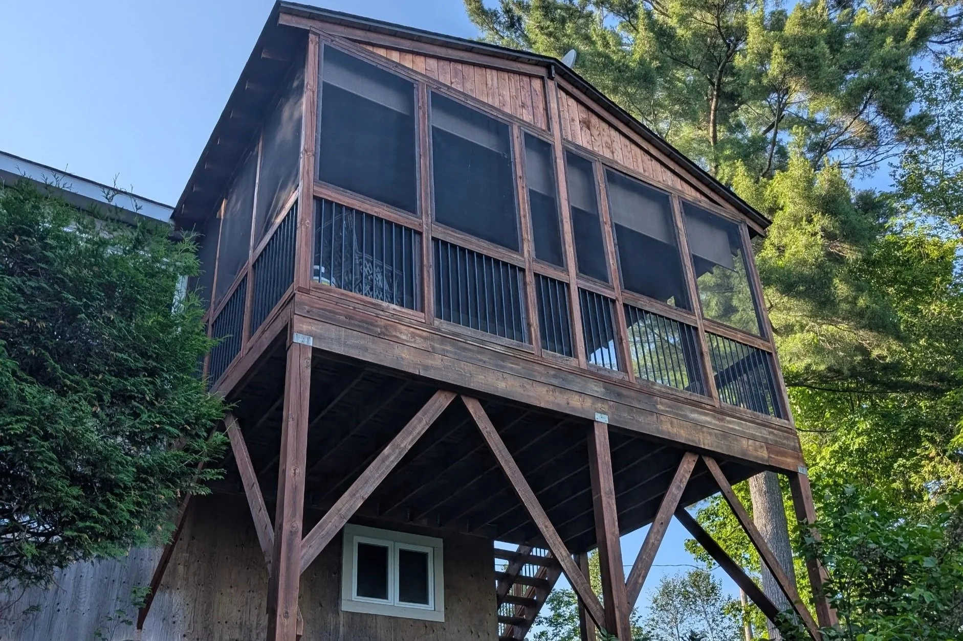 A raised wooden house with screened-in porch, supported by stilts, surrounded by green trees.