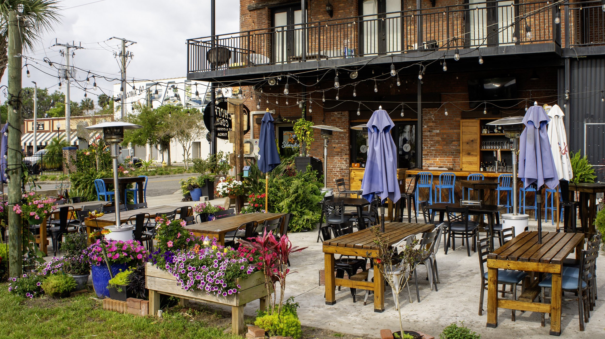 Outdoor seating area of a cafe with wooden tables, blue umbrellas, and string lights. Surrounded by colorful flowers and greenery, the setting provides a cozy atmosphere. The brick building in the background has a balcony and bar area.