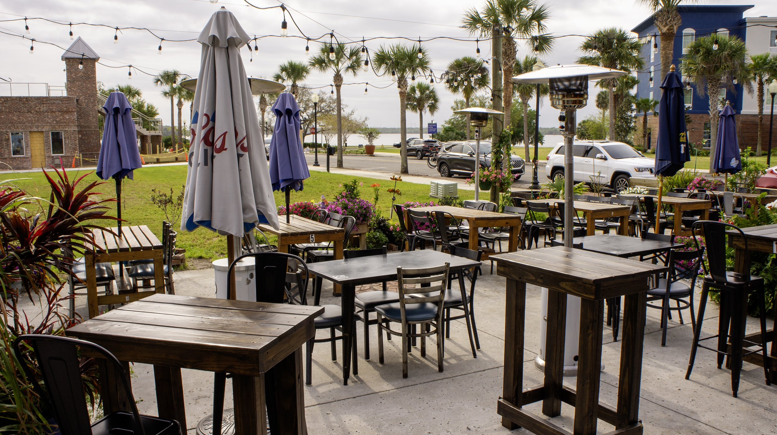 Outdoor patio seating area with wooden tables and chairs, string lights, patio umbrellas, tall heaters, and surrounding palm trees. Cars and a building are visible in the background.