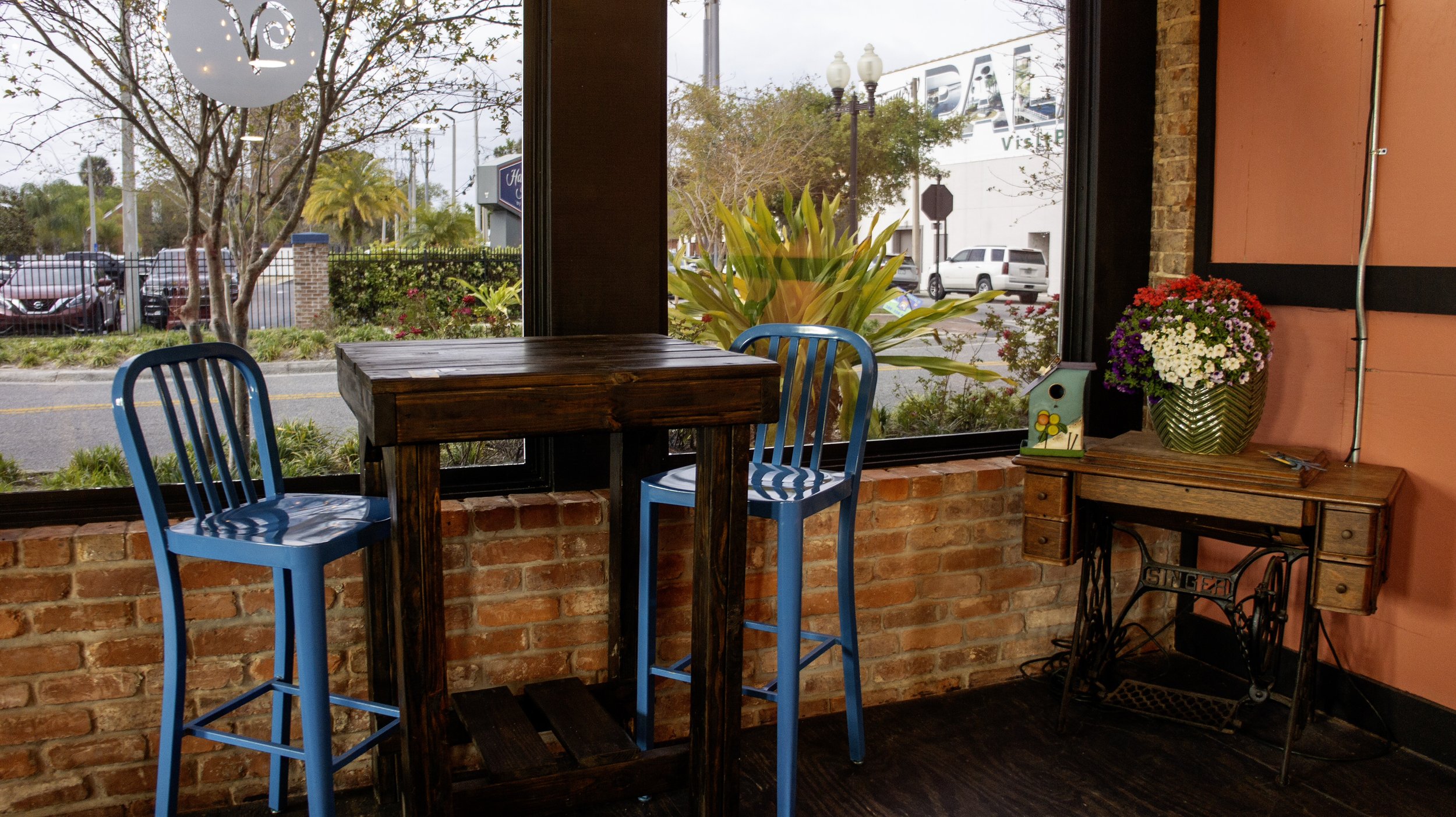 Cafe interior with two blue chairs and a wooden table against a brick wall, next to a sewing machine stand with a floral arrangement and a decorative birdhouse on top.
