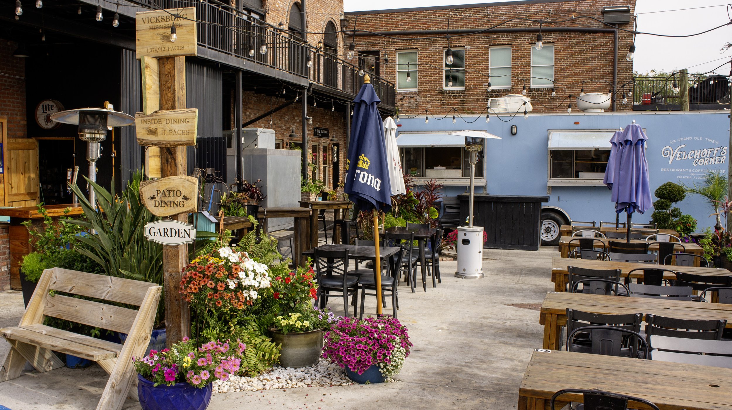Outdoor patio dining area with wooden tables, black chairs, colorful umbrellas, potted flowers, a blue food truck, and a vintage-style brick building in the background.