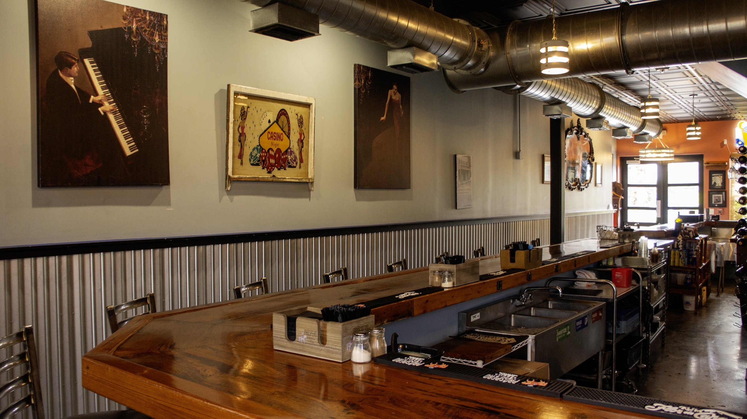 Interior of a bar with a wooden countertop, bar stools, and industrial decor. The walls feature artwork and a "Casino Night" sign. Overhead ductwork and ambient lighting are visible.
