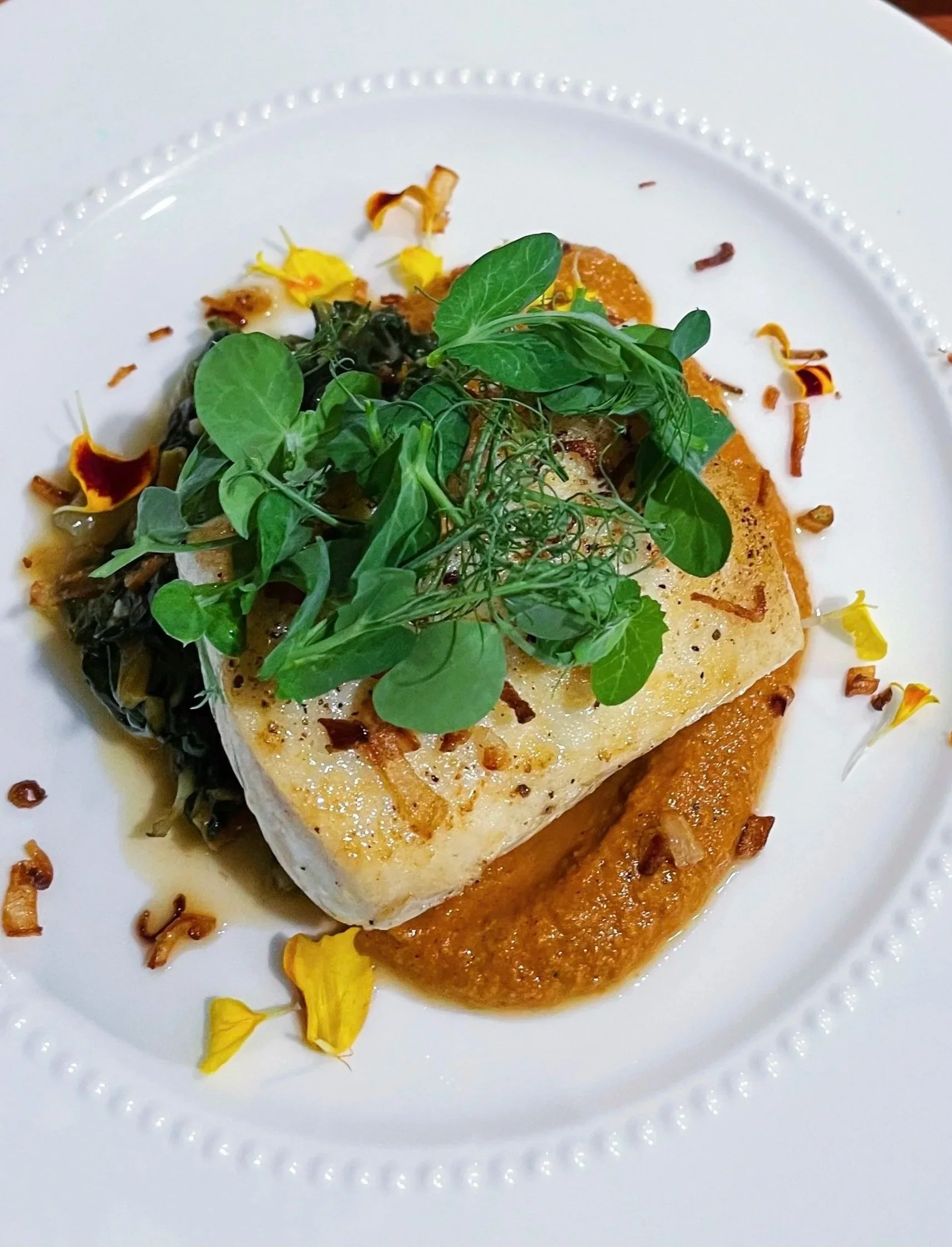 A plated dish featuring a seared piece of fish on a bed of vegetables, garnished with microgreens and edible flower petals, served on a white plate with a decorative border, accompanied by a sauce or puree.