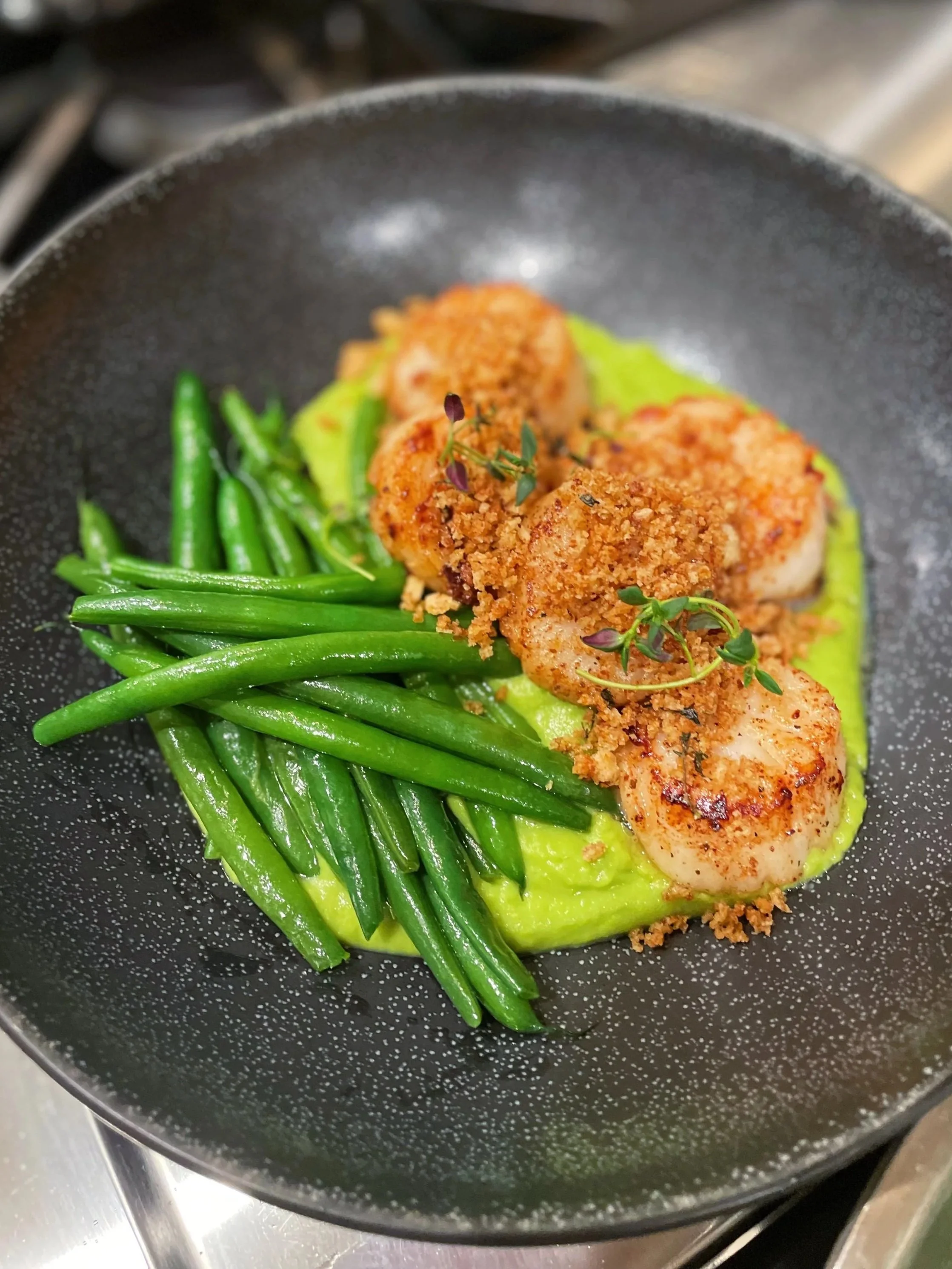 Pan-seared shrimp topped with breadcrumbs served with green beans and a smooth yellow sauce or puree on a black plate.