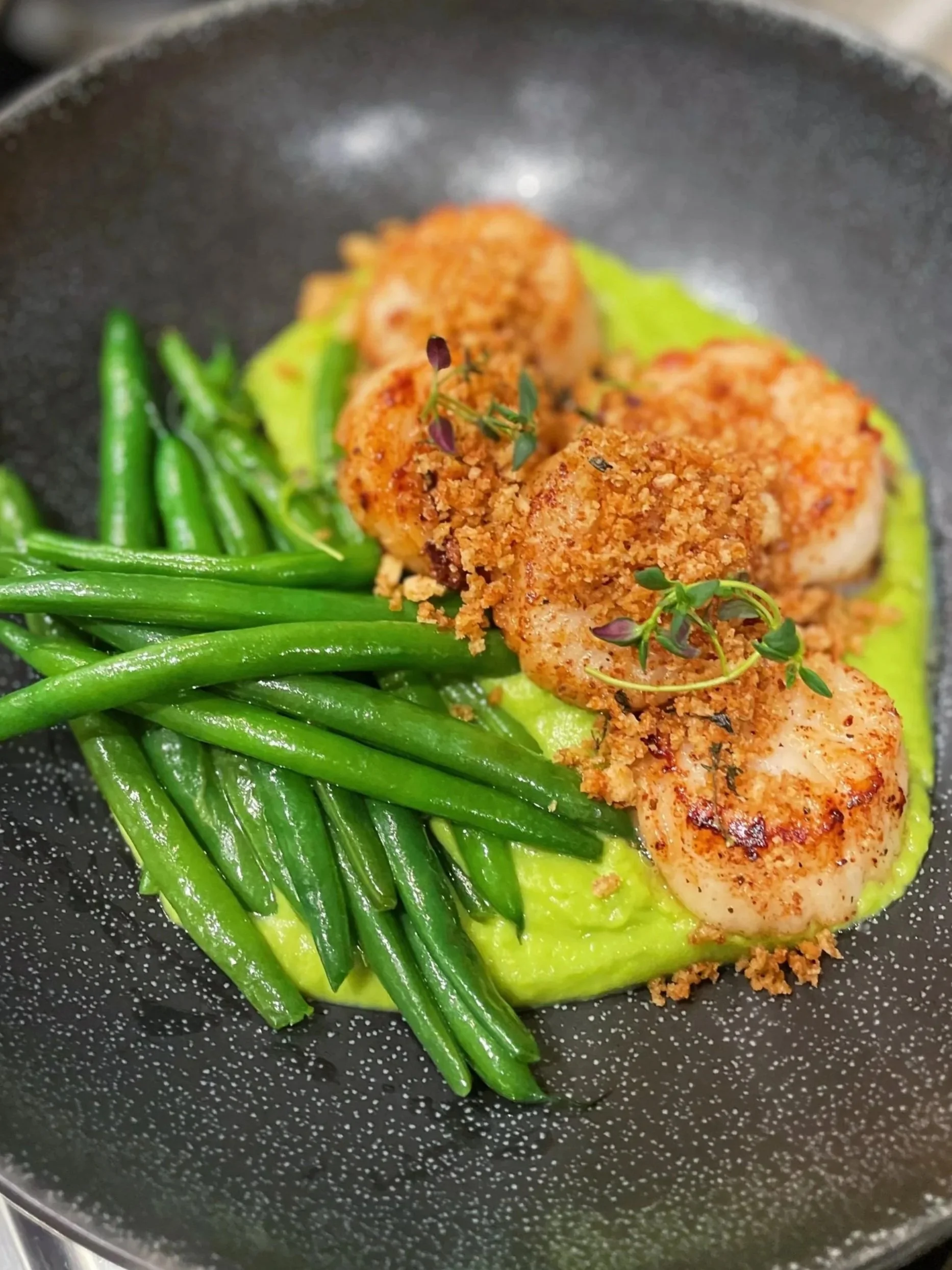 Pan-seared shrimp topped with breadcrumbs served with green beans and a smooth yellow sauce or puree on a black plate.