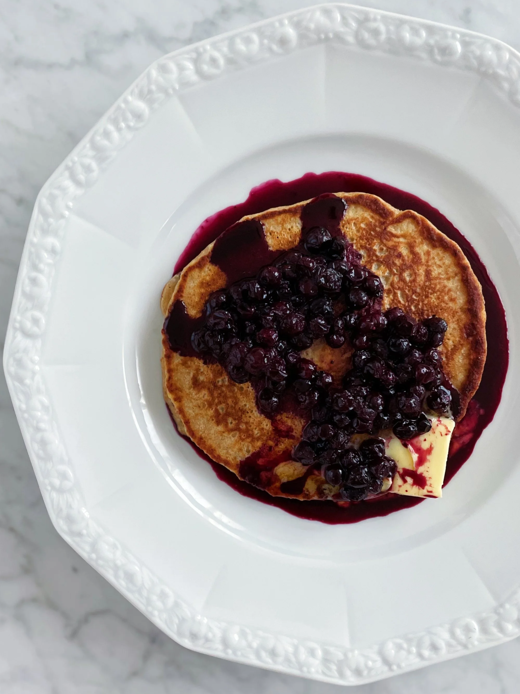 Pancakes topped with blueberries and butter, served with blueberry syrup on a white decorative plate on a marble surface.