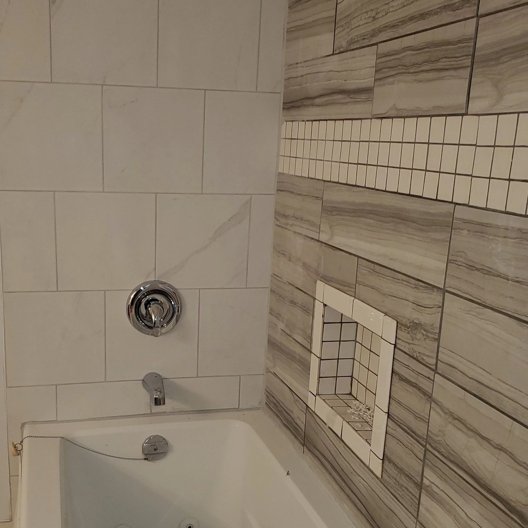 Bathroom corner with modern tile walls, featuring a combination of large white tiles and gray stone-like tiles. A chrome faucet and bath fixture are visible above a white bathtub. A small recessed shelf is embedded in the wall.