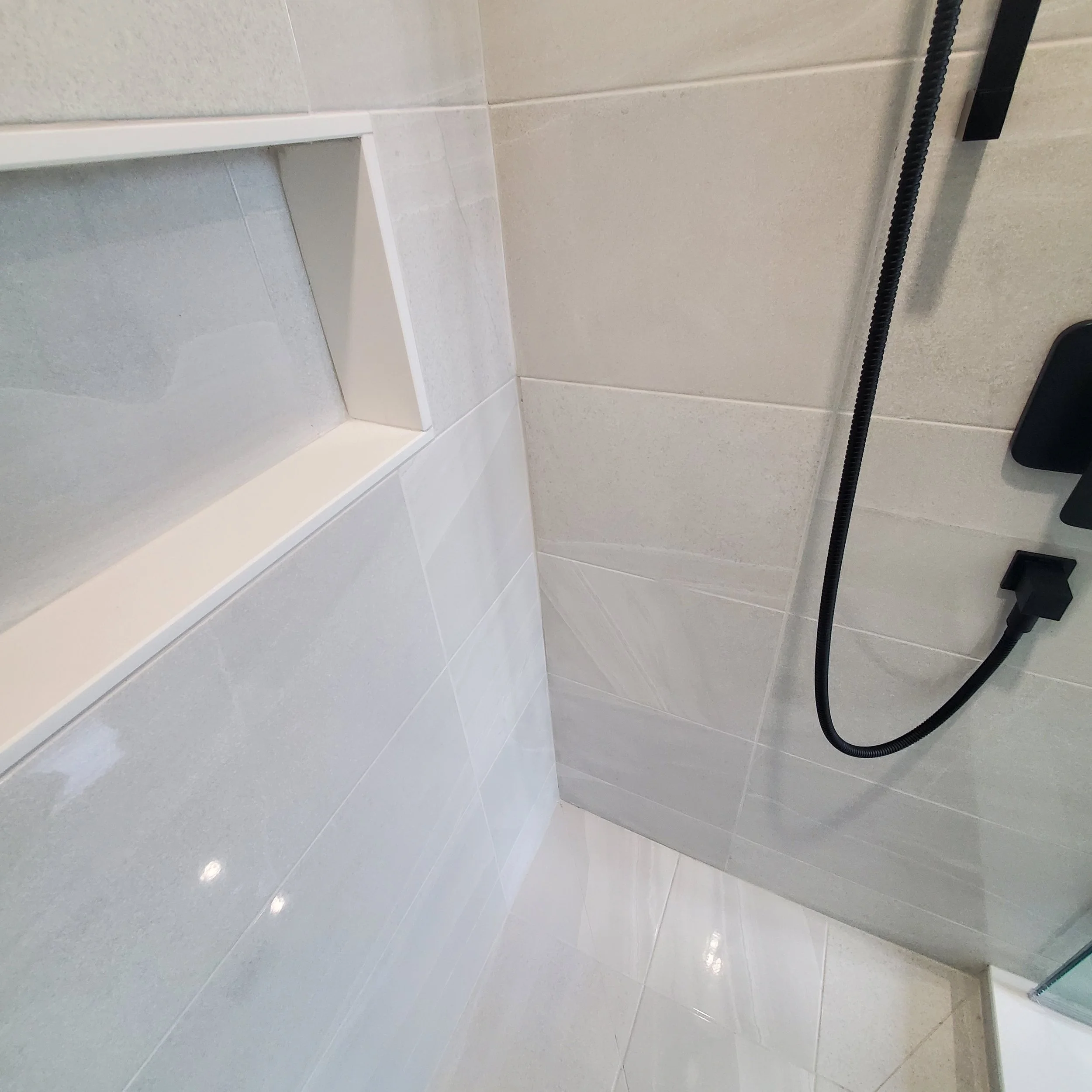 Modern shower with light gray tiles, a recessed shelf, and a black handheld showerhead.