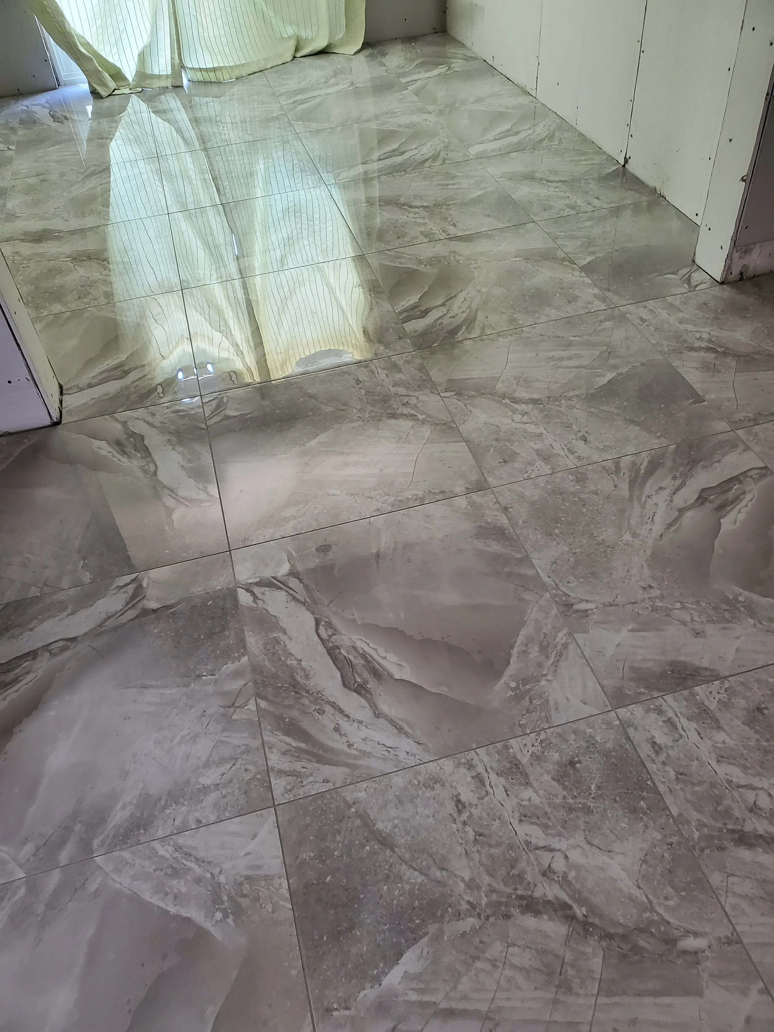 Shiny gray marble tile flooring in a room, reflecting light from a nearby window with curtains.