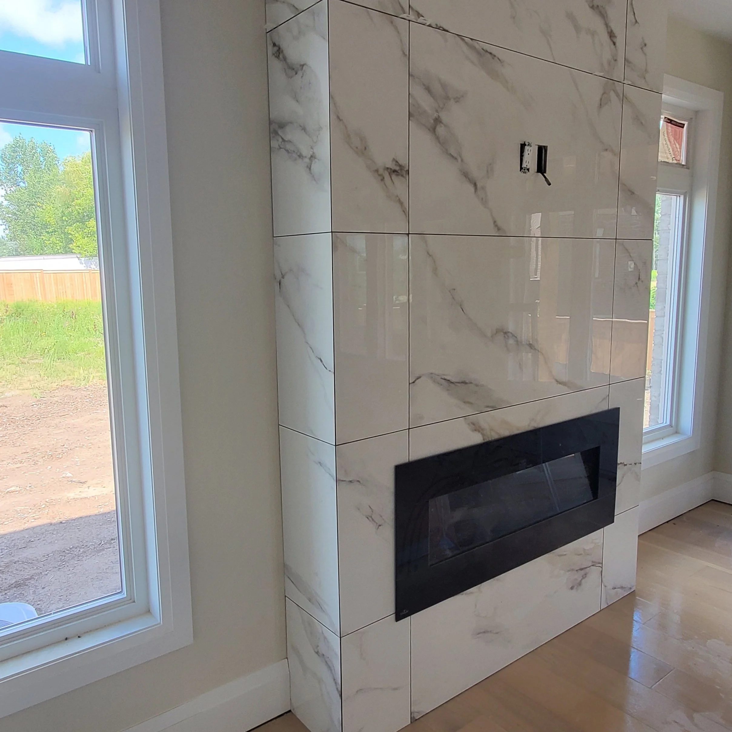 Interior view of a modern fireplace with marble wall tiles and a black inset frame. The room has large windows and wood flooring.