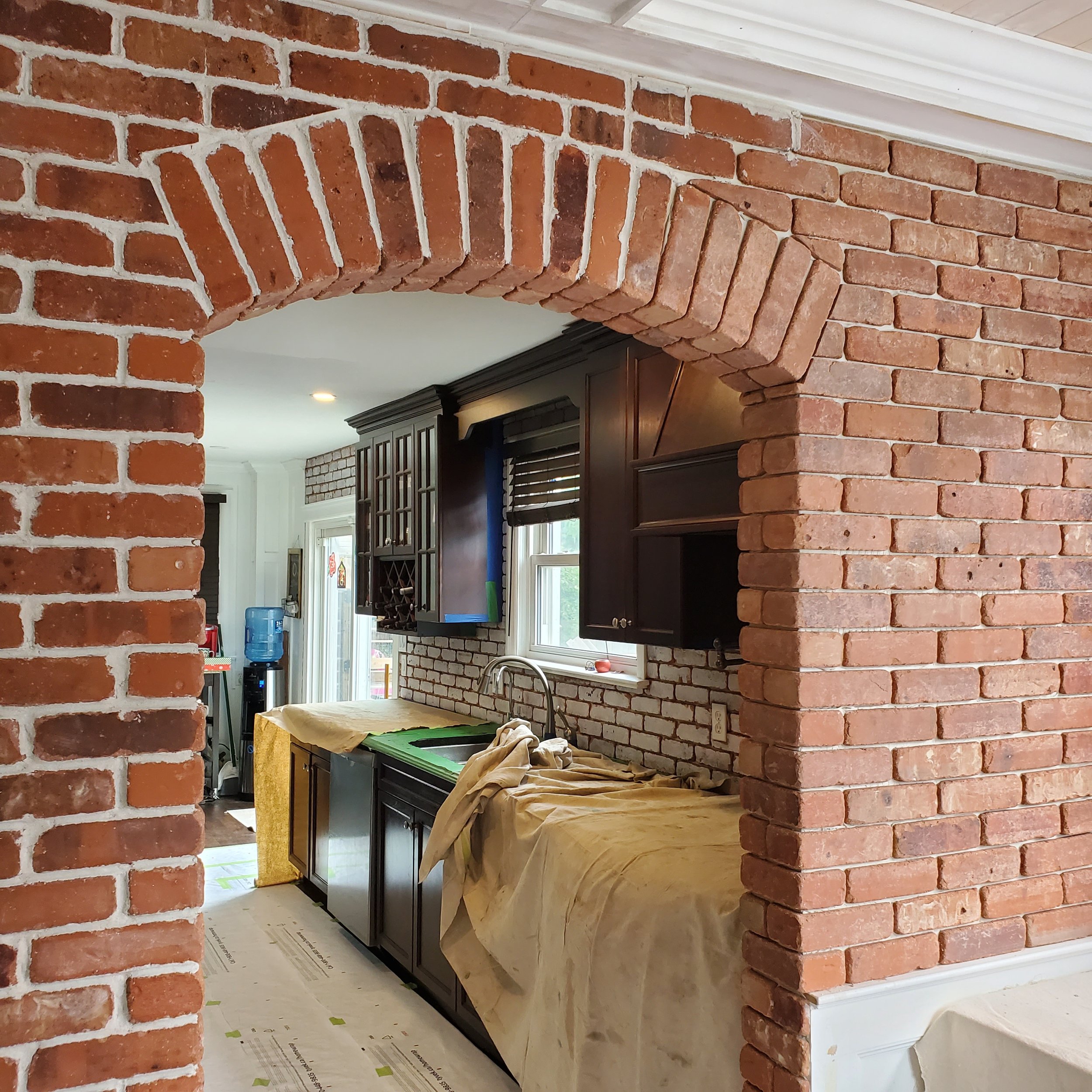 A kitchen under renovation with exposed brick walls, arched brick doorway, and covered countertops. Dark cabinets and a partially visible water cooler are present.