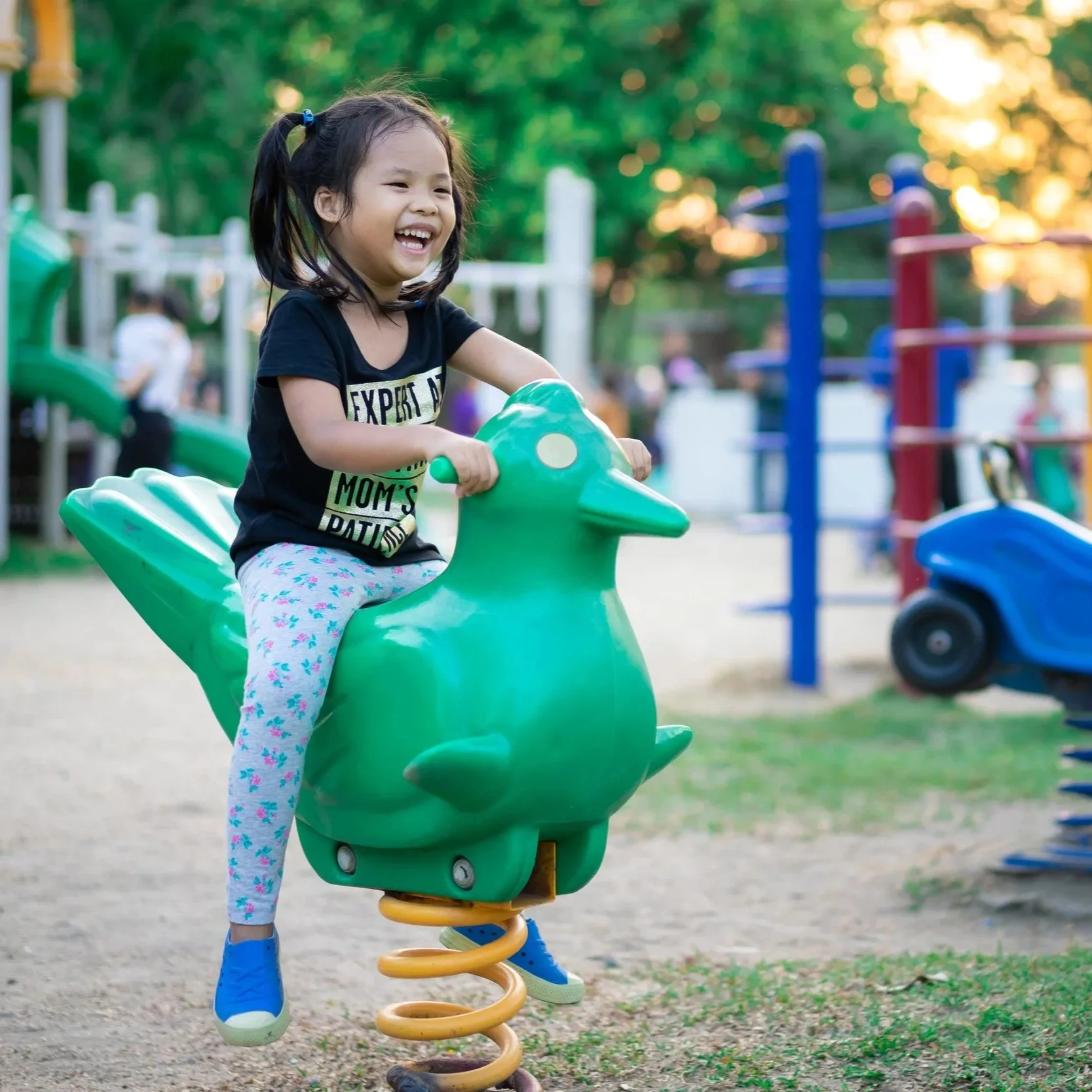 A young child with blonde hair wearing a white shirt sitting on a swing in a park with trees and greenery around, while another child swings nearby.