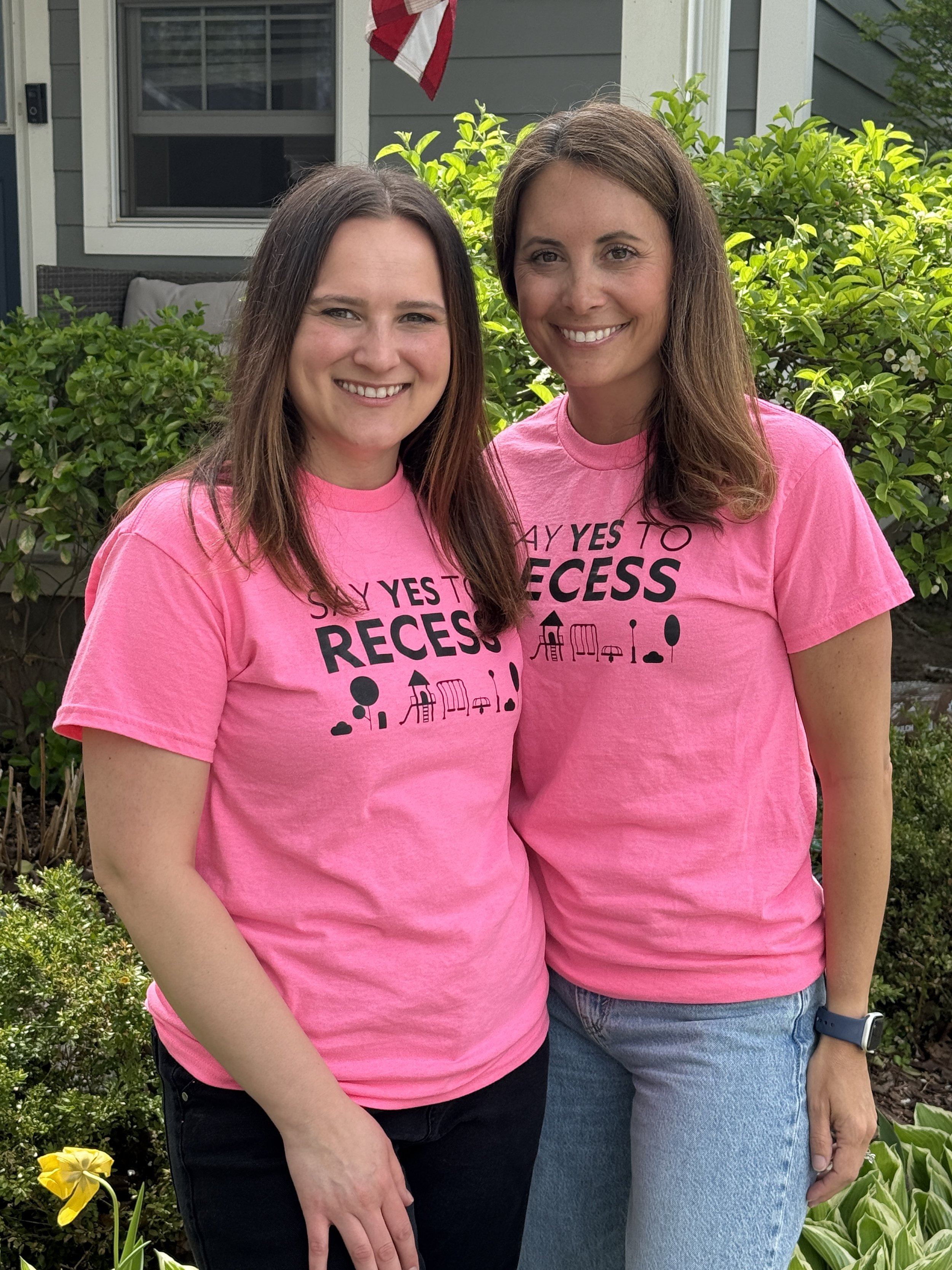 Two women smiling and giving thumbs up while wearing pink T-shirts that say 'YES FOR RECESS' with playground icons, standing outdoors in front of a leafy tree.