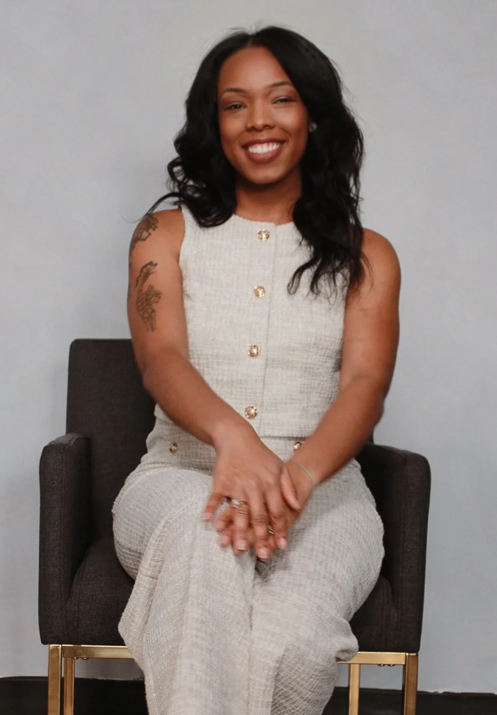 A woman with black wavy hair and tattoos on her left arm, wearing a beige sleeveless buttoned dress, sitting on a dark chair with gold legs, smiling against a plain light gray wall.