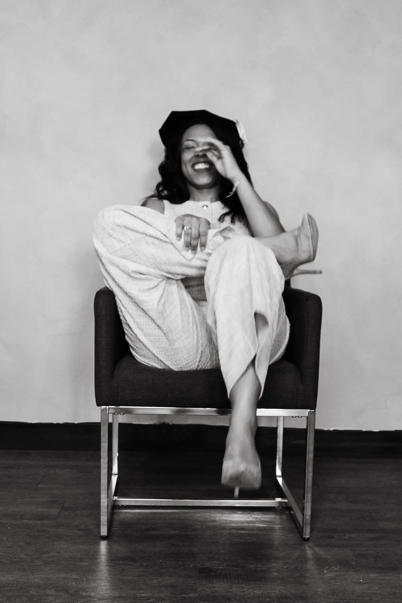 A woman smiling and laughing while sitting on a modern chair against a plain wall, wearing a hat, a light-colored jumpsuit, and high heels.