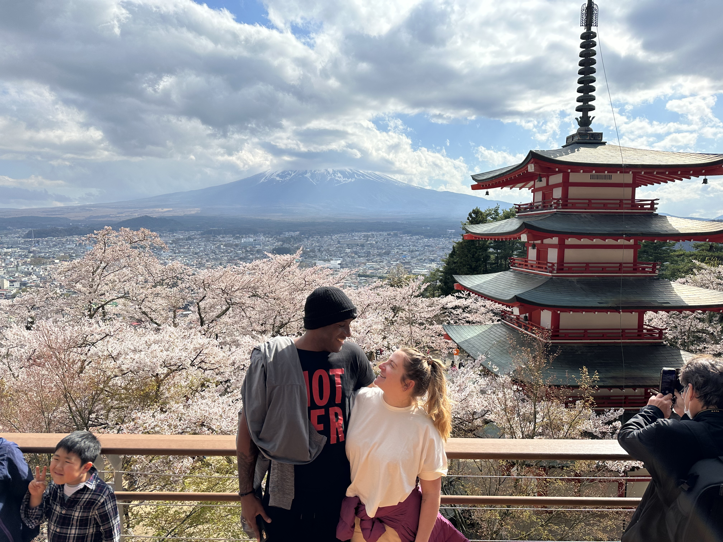 Una pareja sonriente en un mirador con cerezos en flor, en la ciudad con un volcán nevado al fondo, y un pagoda roja a la derecha. Un niño pequeño hace una señal de paz y otra persona toma una foto.
